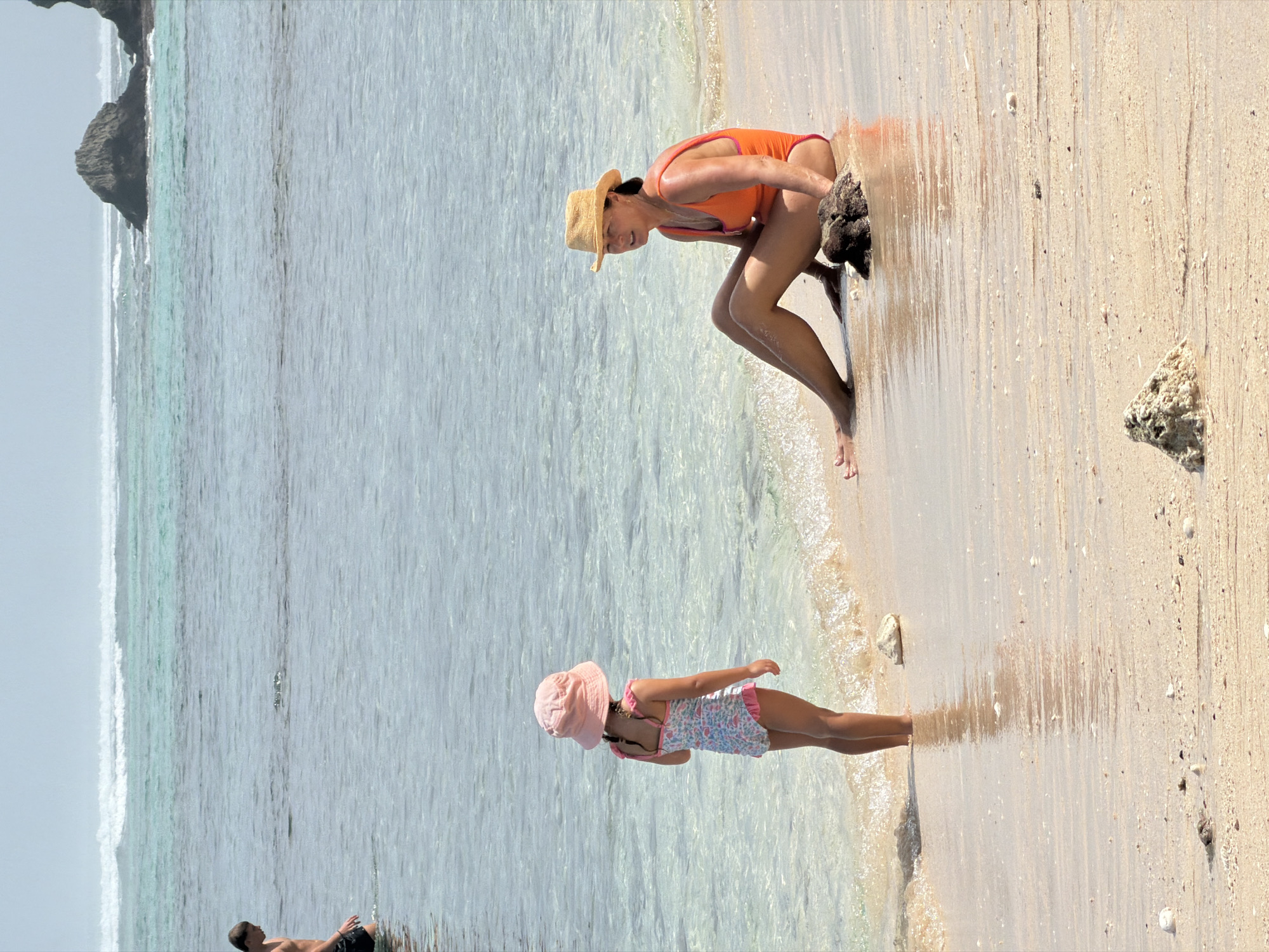 The media file shows a beach scene in Kuta Selatan, Indonesia. The sun is high in the sky, indicating it is likely midday. The weather appears to be sunny and warm, with clear blue water and a sandy beach.
In the foreground, a young girl wearing a pink hat and a floral swimsuit is standing at the edge of the water, her body turned towards the right. She appears to be playing or wading in the shallow water.
Further down the beach, a woman in an orange swimsuit and a straw hat is crouched down, looking towards the water. She is positioned near some rocks on the sand.
In the background, a person is lying on the sand with their back to the camera, facing the ocean. They are wearing dark swim trunks.
The overall scene depicts a relaxed day at the beach with people enjoying the water and the sand. The water is clear and calm, with small waves lapping at the shore. The sand is light brown and appears to be clean. There are a few rocks scattered on the beach, and the ocean extends to the horizon. The image captures a typical tropical beach environment.