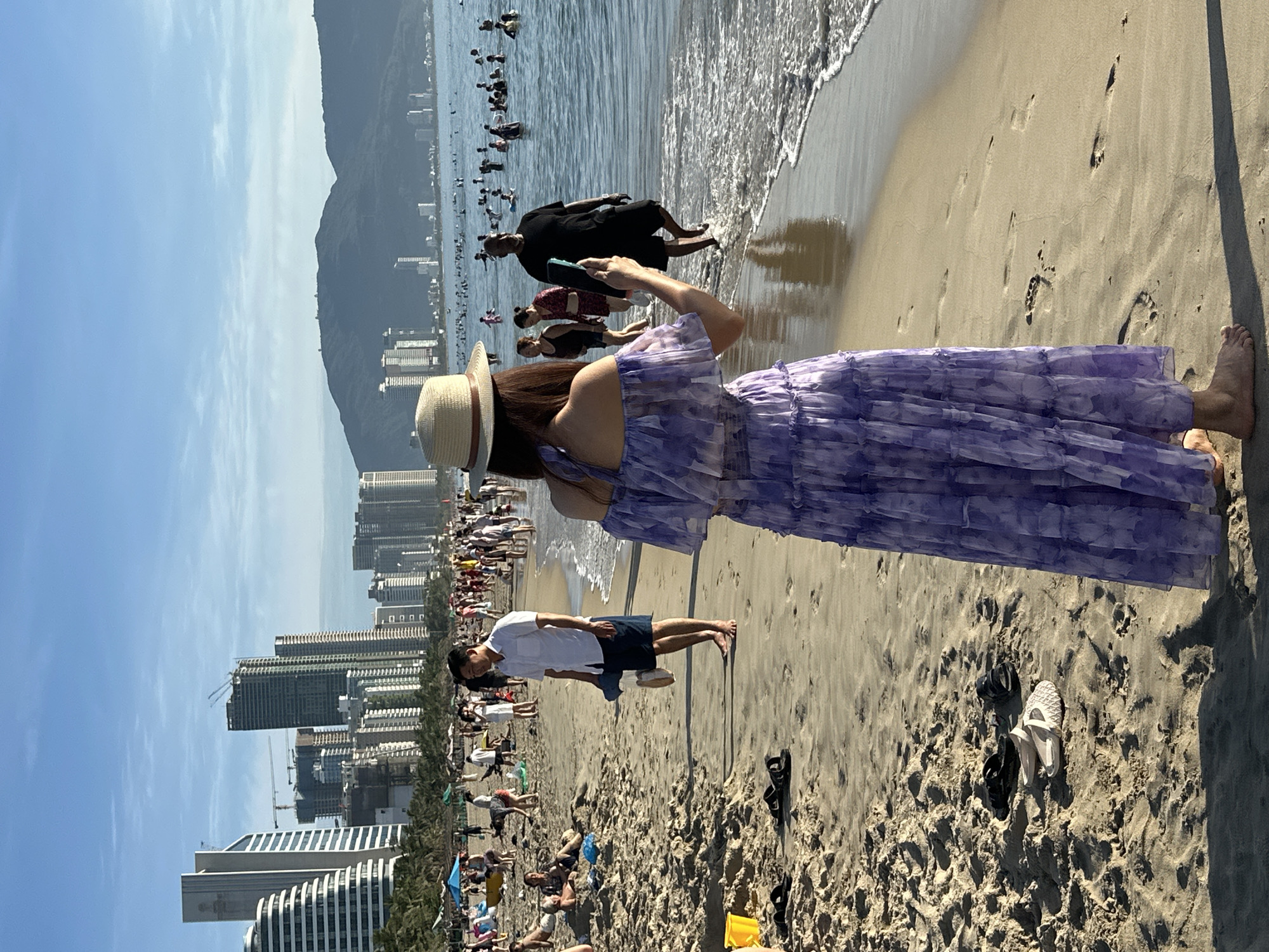 This image depicts a crowded beach scene in Đà Nẵng, Vietnam, likely on My Khe Beach, known for its urban backdrop. In the foreground, a woman wearing a light-colored sun hat and a flowing purple floral-print dress stands with her back to the camera, holding a mobile phone, possibly taking a photo or video. To her left, on the sandy beach, is a pair of black sandals and a pair of light-colored flat shoes. Further back on the beach, a man in a white short-sleeved shirt and dark shorts is walking, carrying a dark bag and a light-colored item. He is barefoot. Numerous people are scattered across the sandy expanse and in the shallow waters of the ocean.
The background reveals the cityscape of Đà Nẵng, with numerous modern high-rise buildings and some under construction, dominating the horizon. Palm trees line the edge of the beach, suggesting a tropical or subtropical climate. The ocean is calm with small waves lapping at the shore, and a large number of people are swimming and wading in the water. The sky is clear with a few wispy clouds, indicating fair weather. The overall impression is a lively and popular tourist destination.