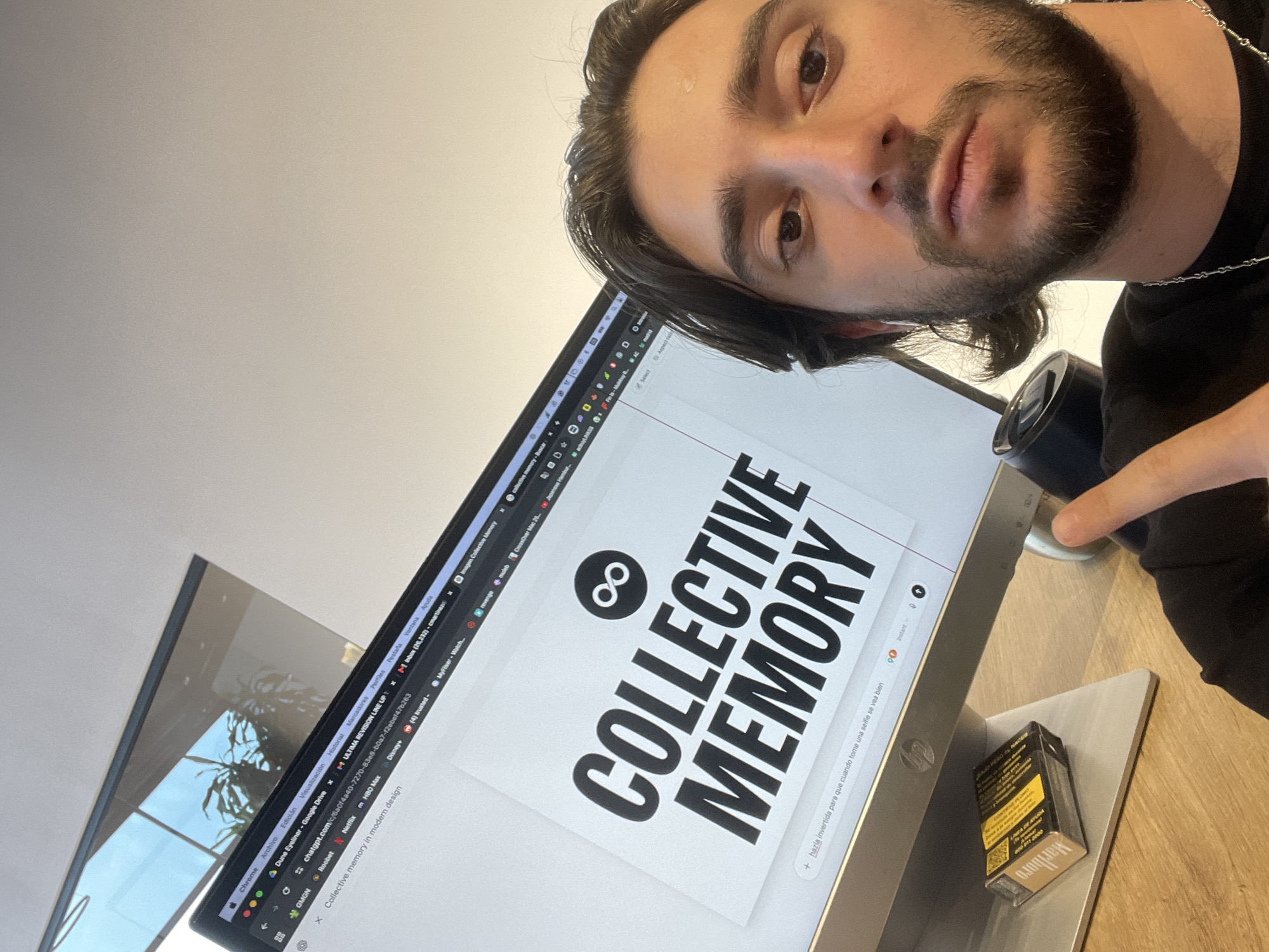 A man with dark hair and a beard is taking a selfie with a computer screen in the background. The screen displays the words "COLLECTIVE MEMORY" with an infinity symbol above. Various browser tabs are visible at the top of the screen, including ChatGPT, Netflix, and Google Drive. A pack of Marlboro cigarettes sits on the desk next to the computer. The location cannot be determined from the image.
