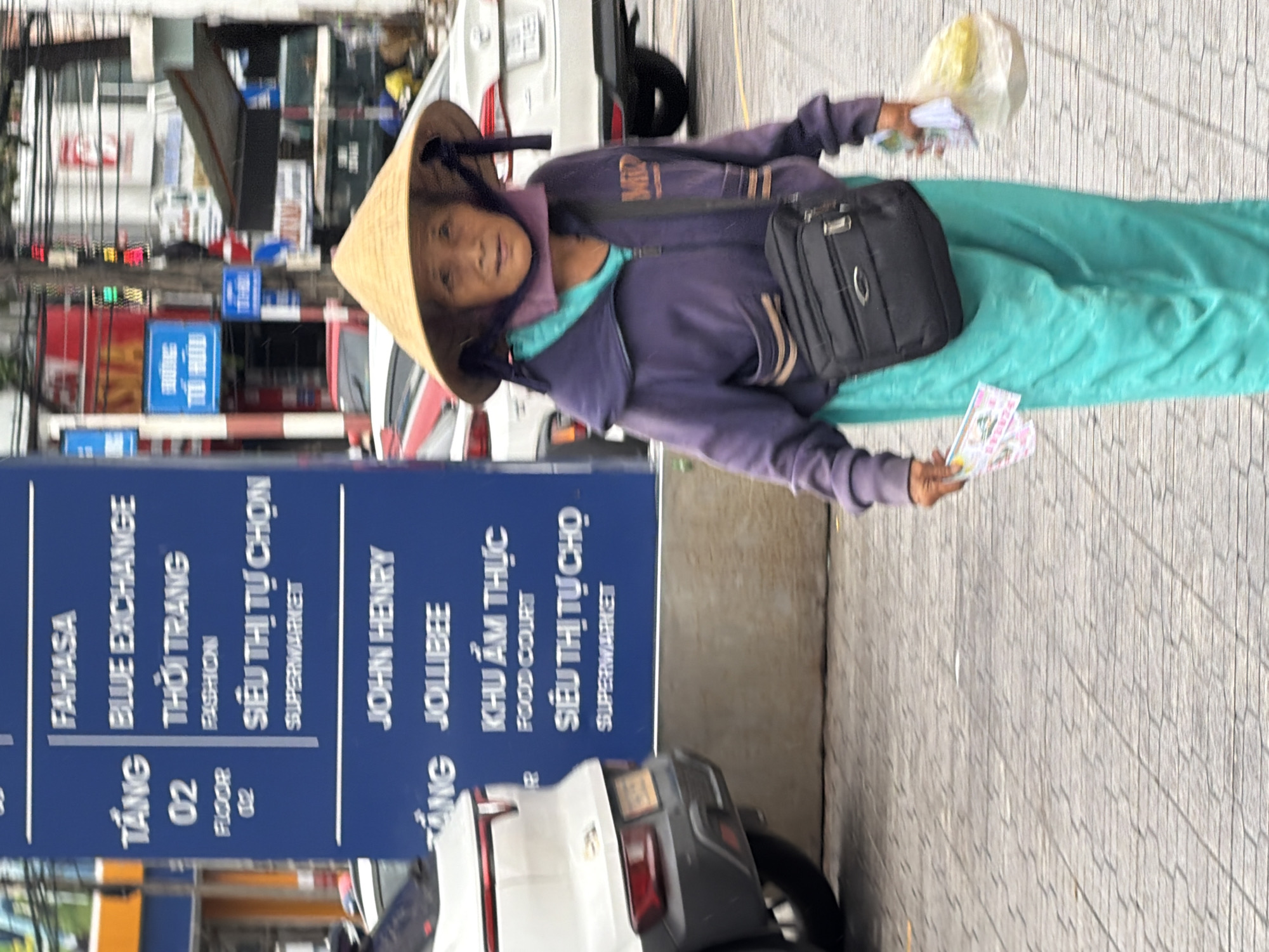 A medium shot captures an elderly Vietnamese woman wearing a conical hat, a purple hooded jacket, and bright teal pants. She is walking on a paved sidewalk in Nha Trang, Vietnam. The woman is carrying a black crossbody bag and holding a plastic bag with what appears to be food in her right hand, and some tickets or coupons in her left hand. Behind her, a blue sign displays text in Vietnamese, listing various businesses and floors, including "FAHASA," "BLUE EXCHANGE," "THỜI TRANG" (Fashion), "SIÊU THỊ TỰ CHỌN" (Supermarket), "JOHN HENRY," and "JOLLIBEE." A white car is visible to the left of the woman, and other vehicles and buildings are blurred in the background, suggesting a busy urban environment. The lighting indicates it is daytime.