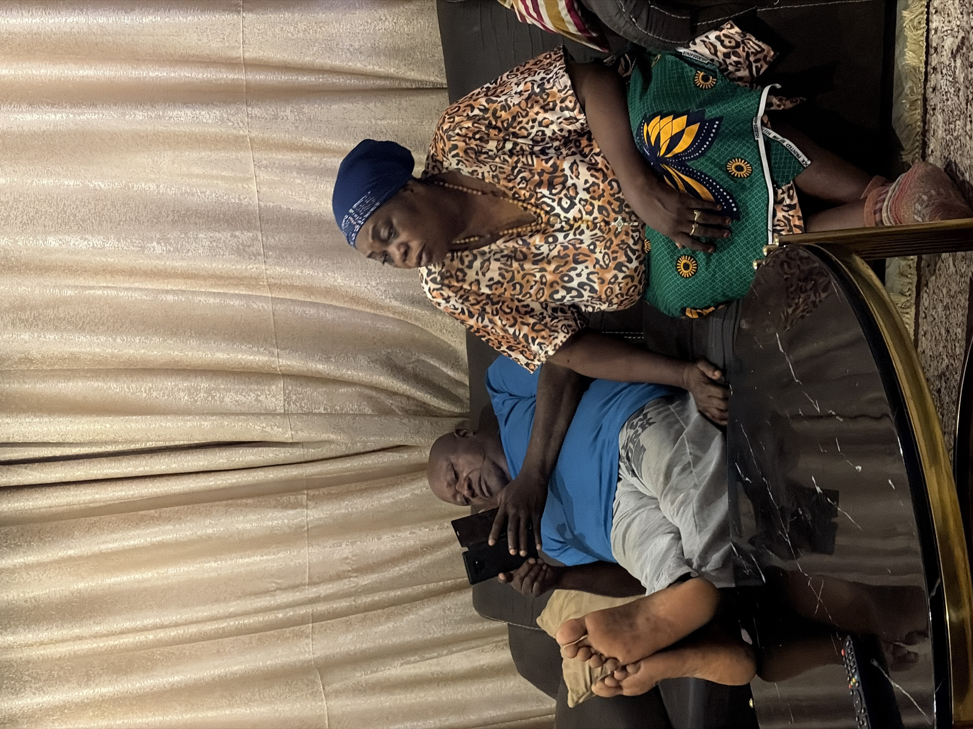 The image depicts a living room scene in Jos, Nigeria. A man, with a bald head and dark skin, is seated on a dark-colored couch. He is wearing a blue t-shirt and grey sweatpants. His bare feet are resting on a black marble coffee table, which also holds a television remote control. The man is holding two dark-colored mobile phones, looking down at them. To his right, a woman is seated on the same couch, facing towards the left of the frame. She is wearing a headscarf, a leopard-print top, and a green and yellow patterned skirt. She has several necklaces on and rings on her left hand. Behind them, beige curtains with a textured pattern hang from a rod. The lighting suggests an indoor setting, possibly in the evening.