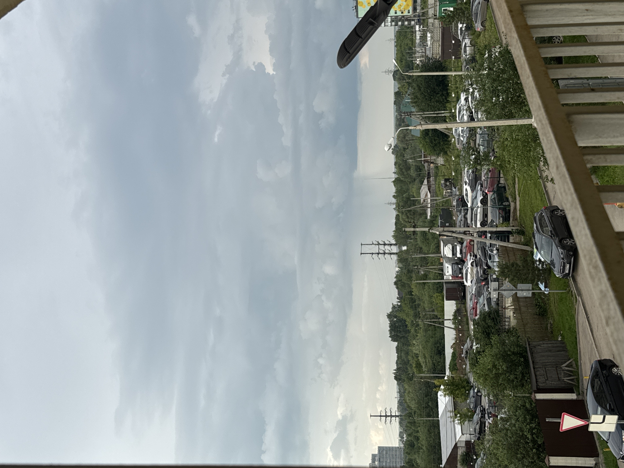 The image shows a view from a balcony overlooking a parking lot and a wooded area, with a cloudy sky above. The scene is set in Kondratovo, Russia. A row of parked cars, mostly sedans and SUVs, are visible in the lot, with some interspersed with trees and bushes. Beyond the parking lot, a dense forest stretches across the landscape. Utility poles with power lines crisscross the scene, indicating the presence of infrastructure. In the foreground, the railing of the balcony is visible, suggesting the photo was taken from an elevated position. There are no people visible in the image. The time of day is difficult to determine precisely due to the overcast sky, but it appears to be daytime. The weather is cloudy and possibly humid, with a soft, diffused light. A street lamp is visible in the middle ground, and a white triangular road sign with a red border is present in the lower right portion of the frame, though the symbol on it is not clearly discernible. The overall mood is somewhat subdued due to the weather and the mundane nature of the scene.