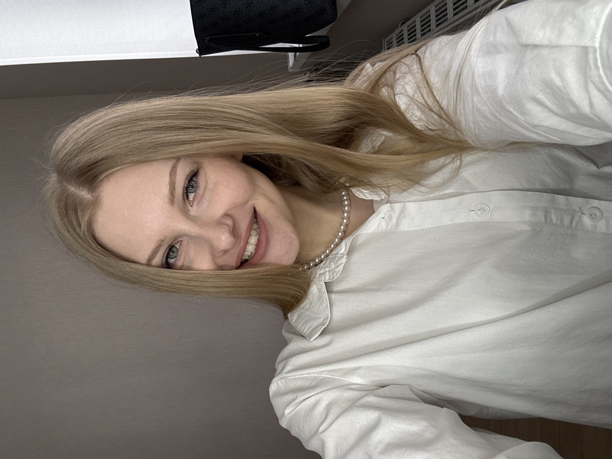 A blonde woman with fair skin is smiling at the camera. She is wearing a white collared shirt and a pearl necklace. The background is a muted grey-brown. There is a Guess brand purse in the upper left corner of the image. The location is not discernible from the image.