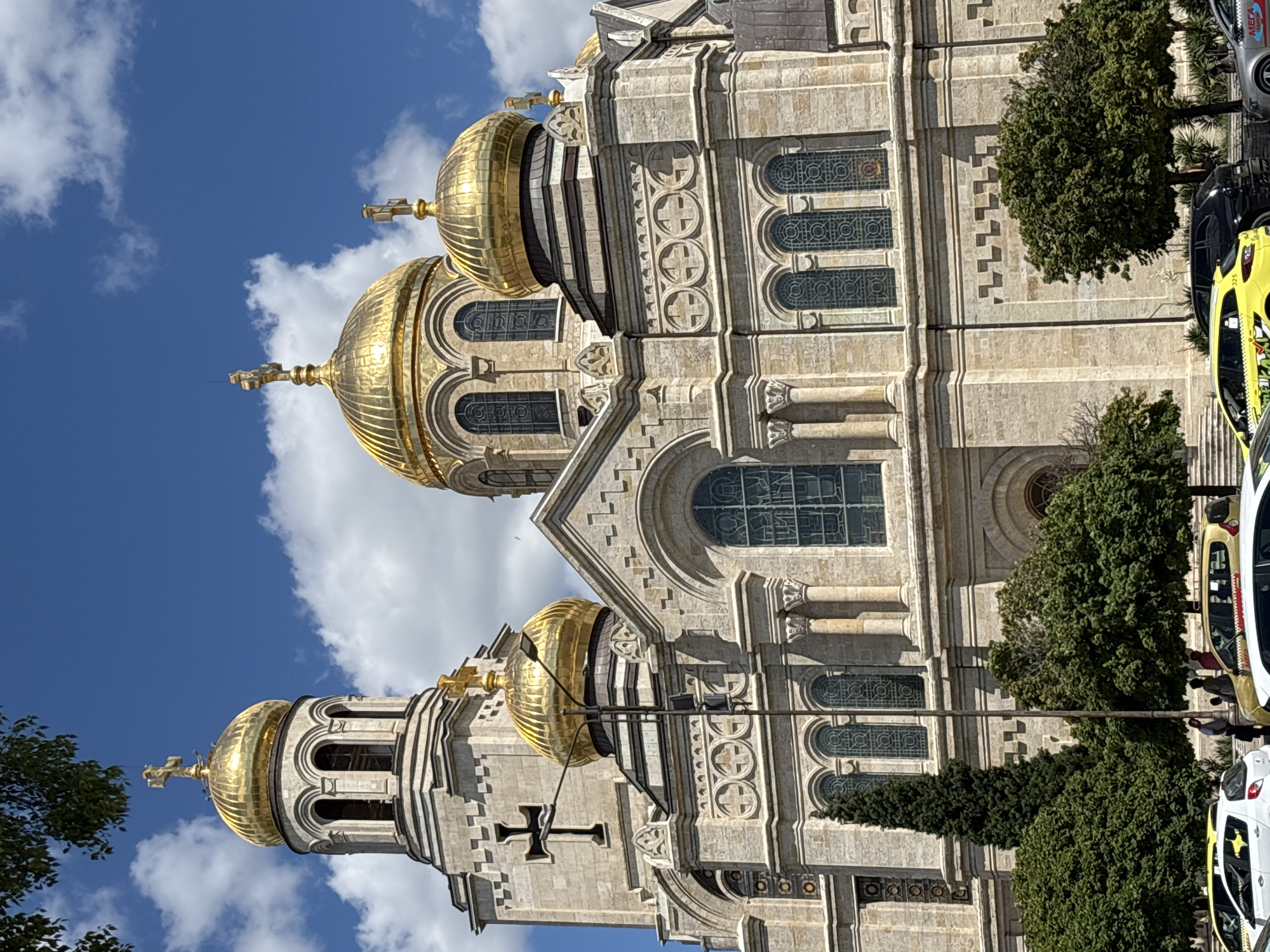 The image shows the Cathedral of the Assumption of the Virgin in Varna, Bulgaria. It is a large Orthodox cathedral with several golden domes and crosses. The facade is made of light-colored stone and features arched windows and intricate carvings. In the foreground, there are trees and several cars, including yellow taxis. The sky is blue with some white clouds. The overall scene depicts a sunny day in front of a prominent religious building.