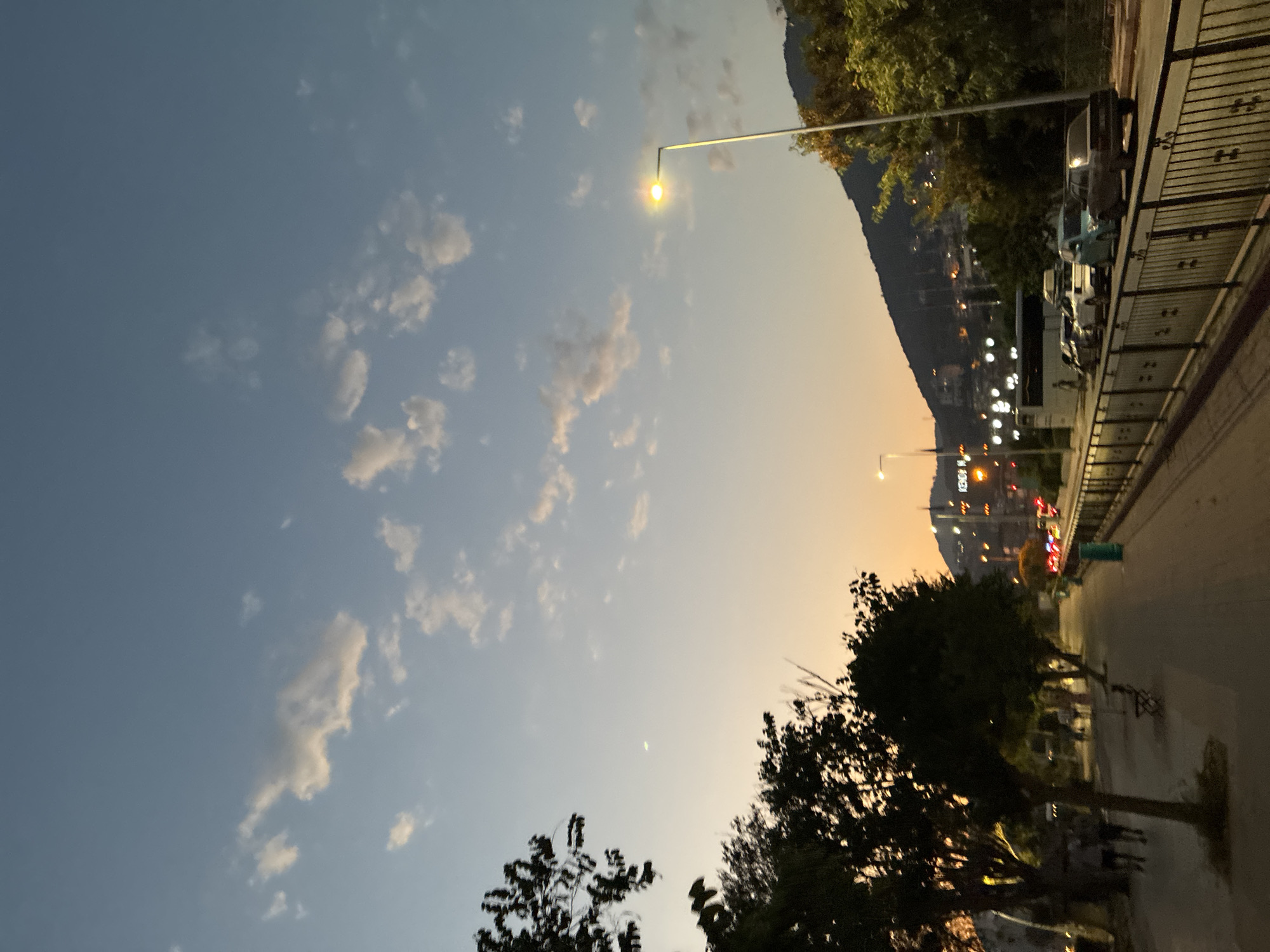 The image captures a coastal street scene in Alanya, Turkey, during twilight. The sky is a gradient of deep blue at the top transitioning to a soft orange and yellow near the horizon, with scattered, wispy clouds. A single streetlamp illuminates the foreground path on the left, casting a warm glow.
To the left, a tree-lined promenade features a paved walkway where several silhouetted figures are casually strolling. Benches are visible along the walkway, one occupied by a person. Trees with dark foliage overhang parts of the path.
On the right side of the image, a road runs parallel to the promenade, separated by a decorative metal railing. Several cars are parked or driving on this road, their headlights and taillights adding points of light to the scene. A large tour bus is also present.
In the background, the city of Alanya sprawls up the side of a dark, mountainous landscape. Numerous lights twinkle across the hillside, indicating buildings and structures. The overall atmosphere is serene and suggests the transition from day to night in a coastal urban environment.