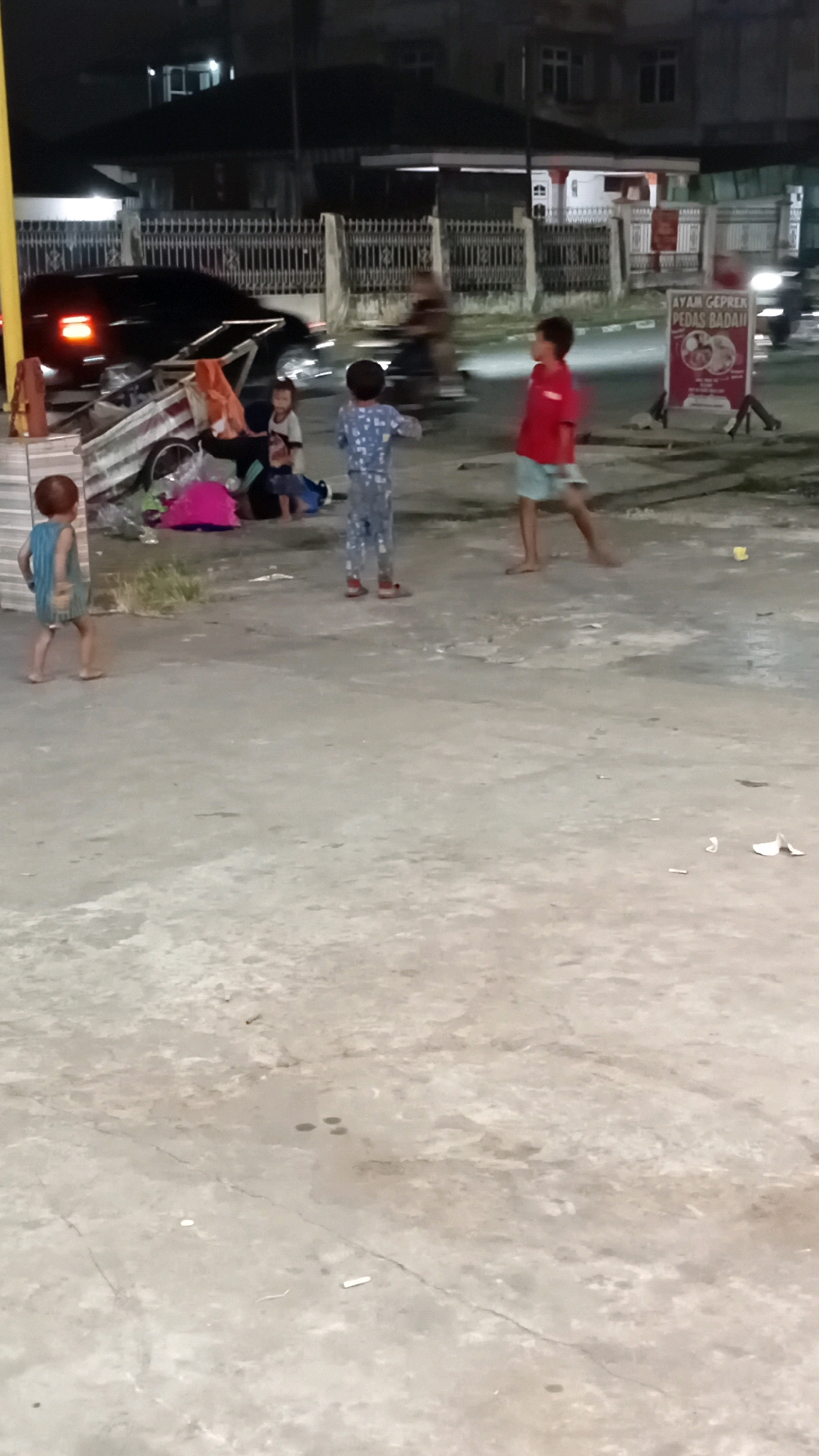The image depicts a nighttime scene on a concrete surface, likely a sidewalk or street, in Jambi, Indonesia. Several children are present. One child, wearing a light blue striped shirt and dark pants, stands on the left side of the frame, facing away from the camera. To the right of this child, a group of children are gathered around a cart laden with items, including a bright pink object and what appears to be fabric. One child in this group is wearing pajamas. Further to the right, another child in a red shirt and light blue shorts is captured in motion, as if playing or running. In the background, a black car with illuminated taillights is partially visible, alongside a fence and buildings. A sign with text, possibly an advertisement, is also present in the background. The overall environment suggests an urban setting at night.