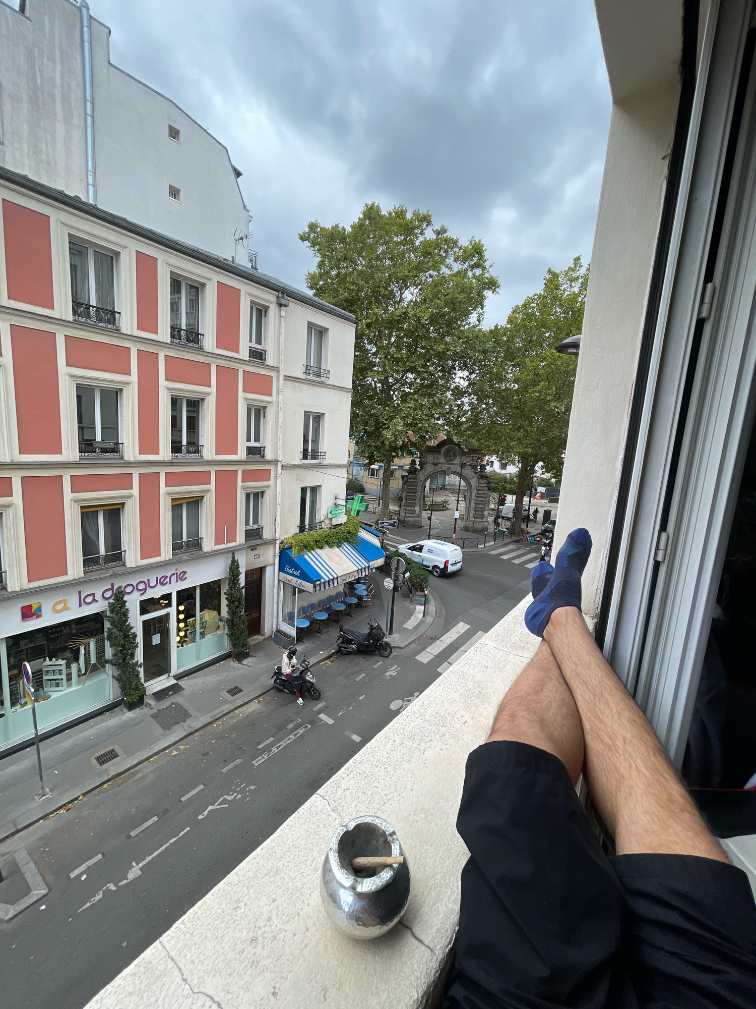 The image shows a street view from an elevated position, likely a balcony or window, in Paris, France. In the foreground, a person's legs are visible, crossed at the ankles and wearing blue socks and black shorts. A metal ashtray with a lit cigarette rests on the ledge.
The scene below depicts a street lined with buildings. On the left, a building with a salmon-pink facade and white trim features multiple windows. A storefront below, labeled "a la droguerie," has large glass doors and displays merchandise. To the right of this building, a street with parked motorcycles and a scooter is visible. Across the street, a building with blue and white striped awnings covers outdoor seating for a cafe. Further down the street, a white van is parked, and an arched gateway with ornate stonework is visible in the distance. Trees with lush green foliage line the street and border the buildings. The sky is overcast with gray clouds.