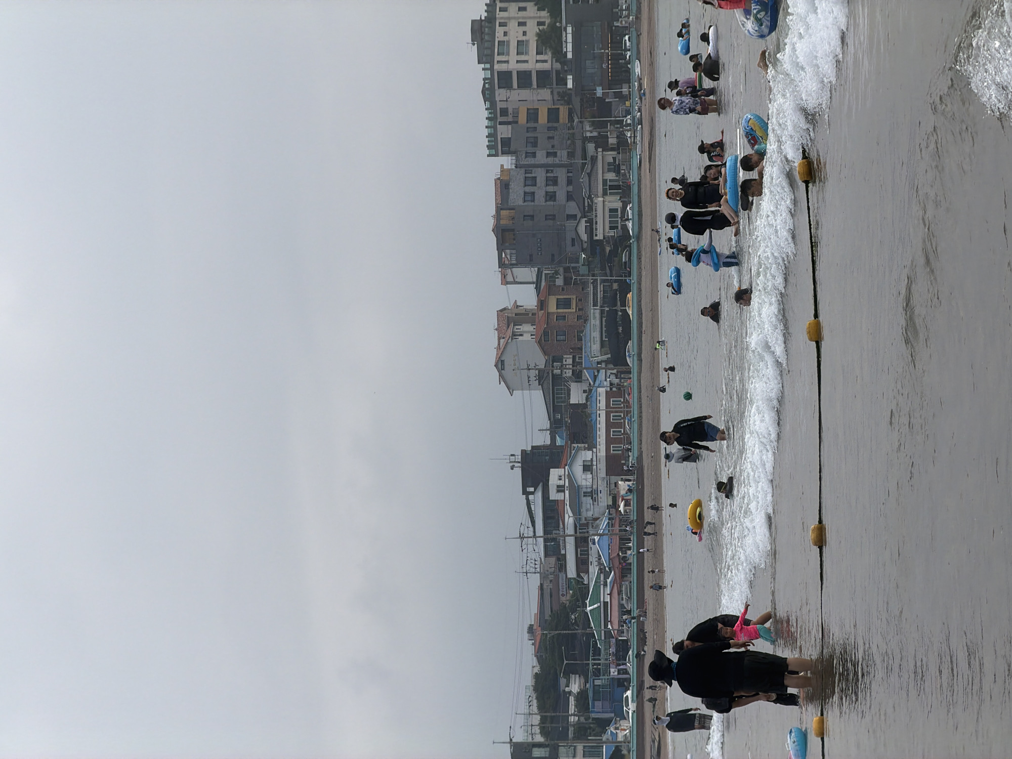This is a medium-shot, eye-level, vertical photograph of a crowded beach in Jeju City, South Korea, on an overcast day. The foreground is dominated by the ocean, with gentle waves lapping at the shore. Several people are visible in the water, many of them children, some playing with inflatable rings and tubes. Yellow buoys and a rope mark a boundary in the water. In the mid-ground, a sandy beach is populated with more people, some wading and others standing. The background reveals a dense urban landscape with a variety of buildings, including multi-story residential structures, smaller shops, and commercial establishments. Power lines and utility poles are also visible. The overall atmosphere suggests a popular public beach on a cloudy day, with recreational activities in full swing.