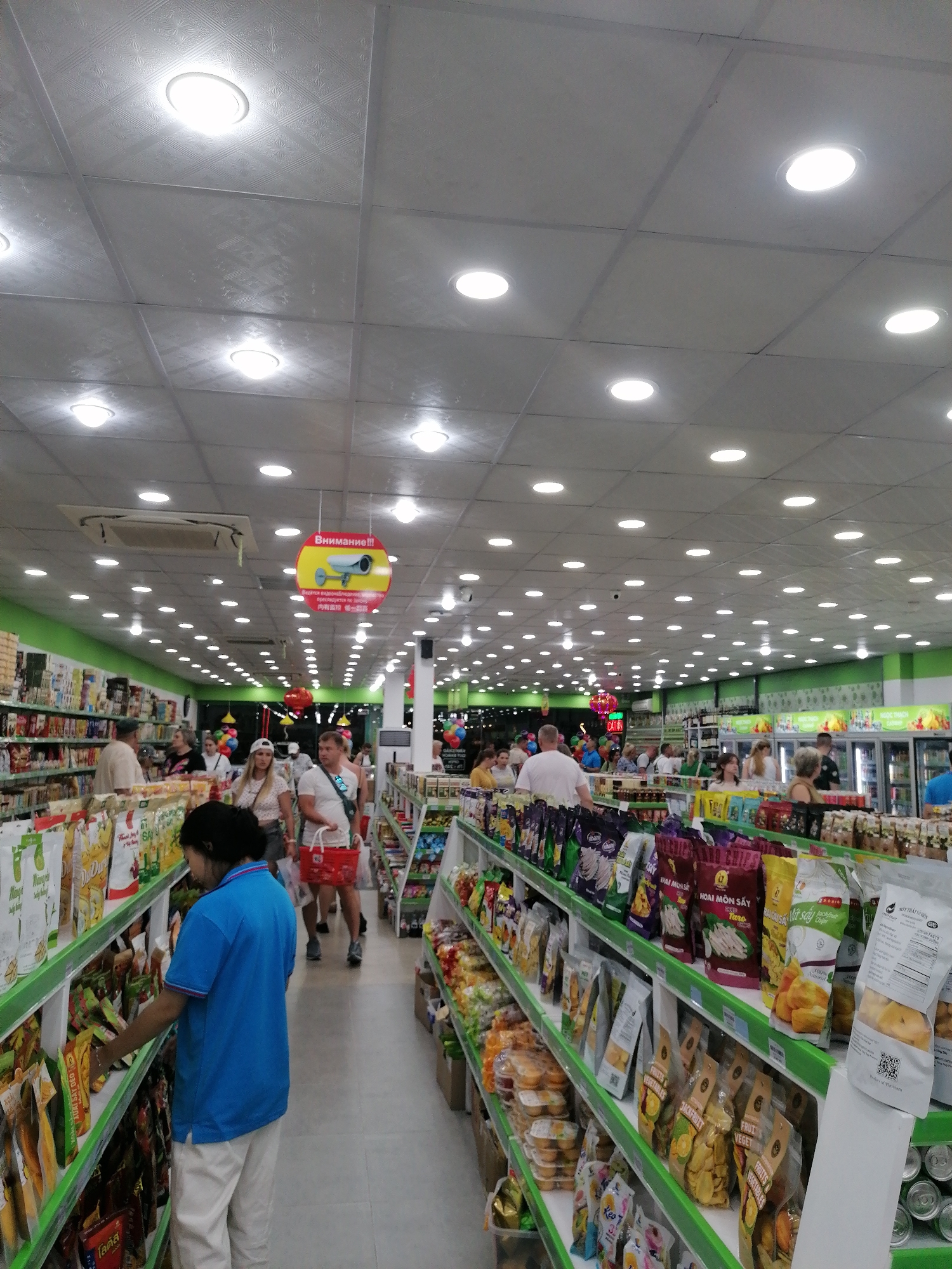 The image shows the interior of a grocery store in Nha Trang Ward, Vietnam. The store is brightly lit with numerous overhead lights arranged in a grid pattern. Shelves lined with various packaged goods, including dried fruits and snacks, run along the aisles. Customers are present, browsing and shopping. A store employee wearing a blue shirt and white pants stands near the left aisle, examining items on the shelf. Signage in Vietnamese and Russian hangs above, displaying notices or warnings. The store is moderately crowded, and the shelves are well-stocked.