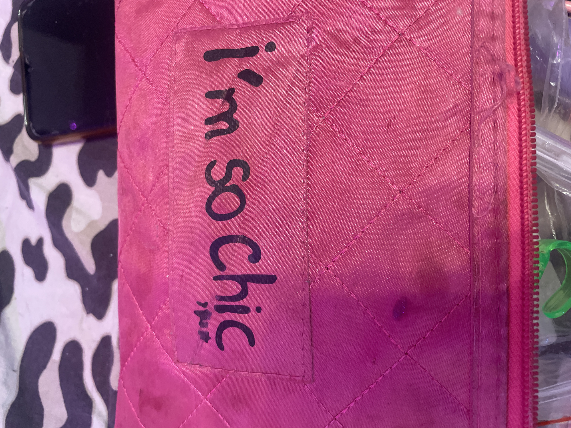 The image shows a close-up of a pink quilted fabric surface, possibly a makeup bag or a similar accessory. There is a clear plastic patch adhered to the fabric, on which the words "I'm so chic" are written in black marker. The writing is somewhat smudged, and there are some dark spots on the fabric itself, possibly stains. The background of the image is a blurry pattern of black and white shapes, resembling a cow print or camouflage. A dark-colored smartphone is partially visible in the upper right corner of the image, resting on the patterned background. The bottom of the image reveals a pink zipper and the edge of what appears to be a clear plastic bag containing various items. The scene is indoors, and the lighting is somewhat dim, casting a pinkish hue over the fabric. There is no indication of the city of Sinyavino, Russia, in the image.
