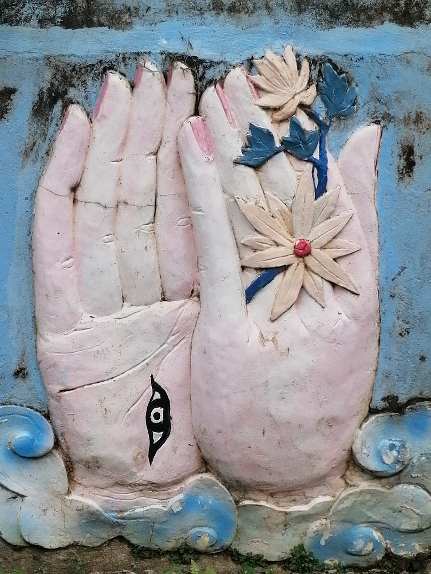 The image displays a bas-relief sculpture of two hands, positioned as if in prayer or offering. The hands are painted in a pale pinkish hue with lighter highlights and darker pink on the fingernails. The left hand is shown palm-up, while the right hand is held palm-down, with the fingers of both hands slightly curled. A decorative element consisting of a stylized flower and leaves is attached to the right hand. The flower is a light cream color with a red center, and the leaves are a blue-green. The sculpture is set against a textured background that appears to be a weathered wall, painted in a faded blue color with dark, grimy streaks. At the base of the hands, there are stylized representations of clouds, painted in blue and white. On the palm of the left hand, there is a black, eye-like symbol with a white iris. The overall impression is that of an outdoor decoration or religious icon. The sculpture is likely located at the Dinh Thu Ong (Ong Temple), also known as Quan De Temple, in Da Lat, Vietnam.