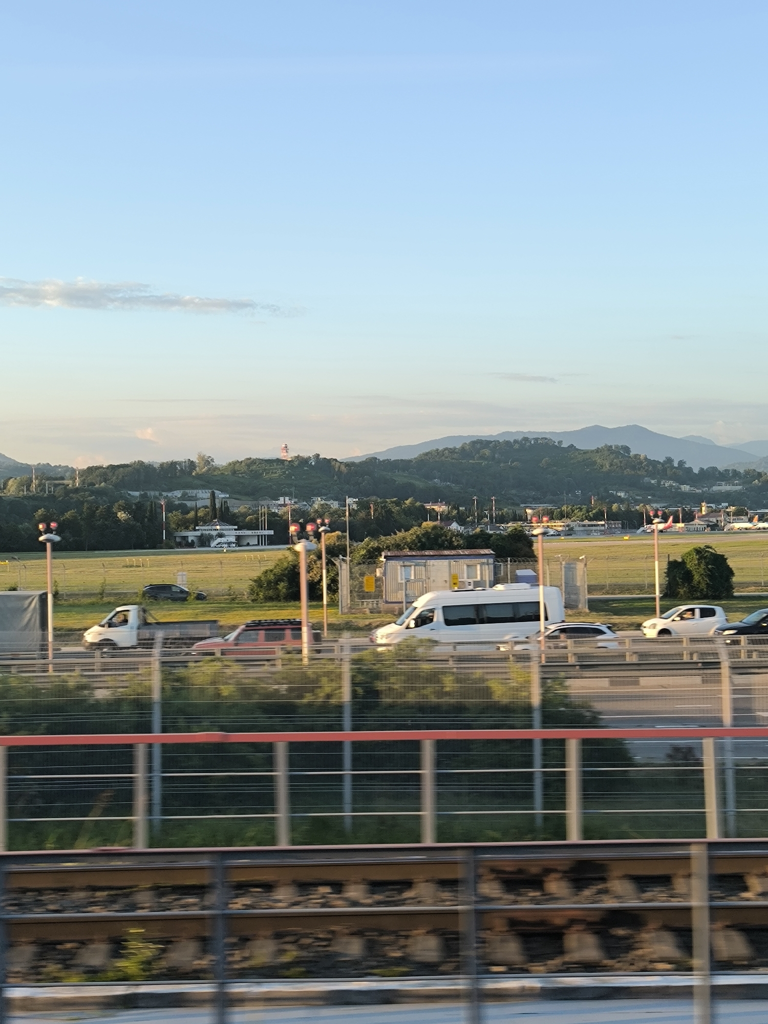 The image shows a scenic view of a highway and a nearby airport at sunset. In the foreground, a section of railway tracks with ballast and sleepers is visible. Above the tracks, a red and white metal fence with vertical supports runs horizontally. Further up, a highway with multiple lanes of traffic is depicted, with several vehicles, including cars, a white van, and a pickup truck. The vehicles are in motion, suggesting a dynamic scene. Beyond the highway, an airport runway stretches across a grassy field. Several small buildings, possibly airport facilities, are present in the middle ground. In the background, rolling hills covered in lush green trees create a natural backdrop. A faint tower with red lights is visible on one of the hills. The sky is a gradient of light blue, with a few wispy clouds near the horizon. The overall lighting suggests the warm tones of a late afternoon or early evening sun.