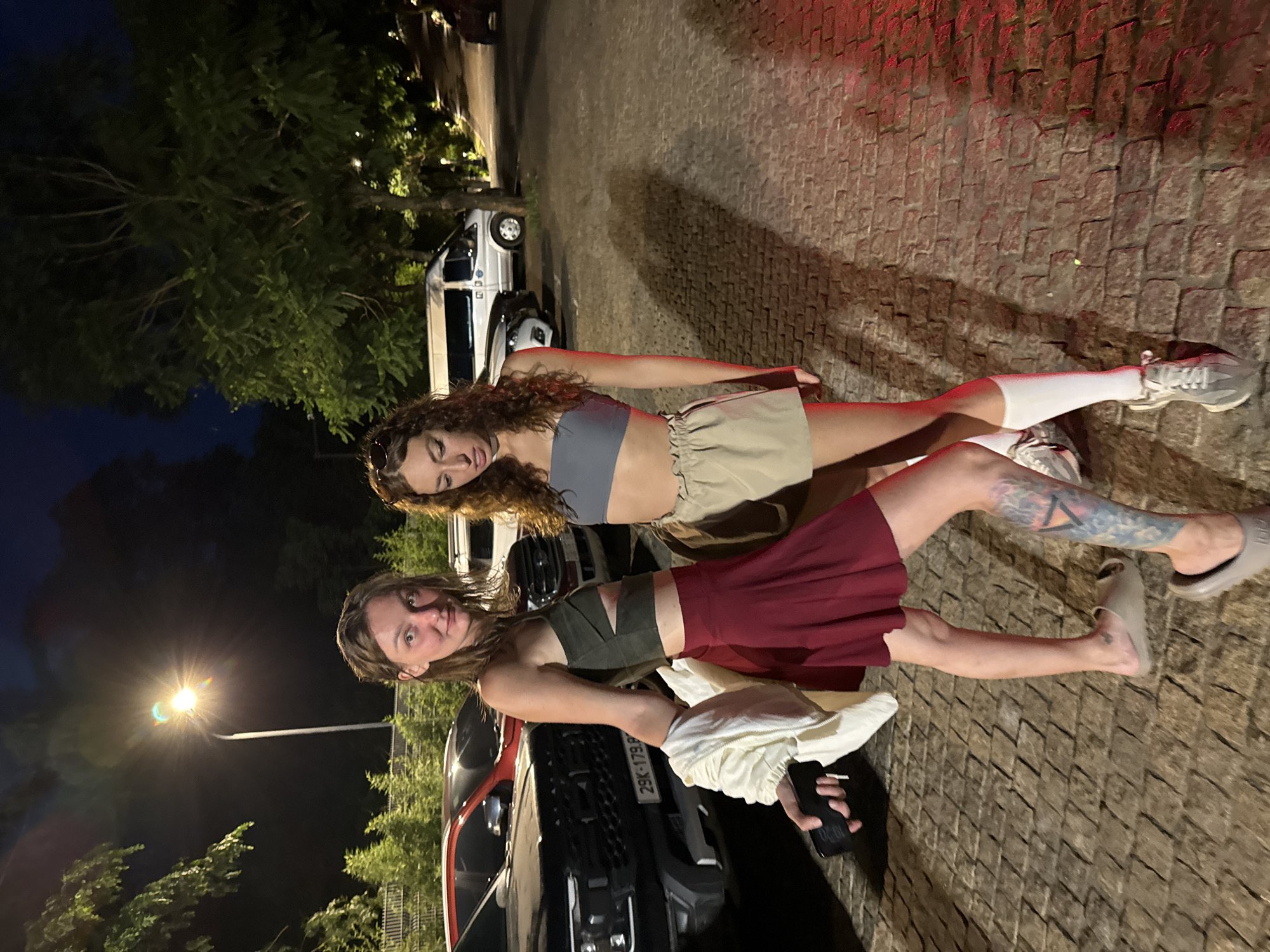 The image, when correctly oriented, features two young women standing on a textured paved surface at night, likely in Púng Luông, Vietnam.
The woman in the foreground (left side when oriented correctly) has medium-length brown hair and a slight smile directed towards the camera. She wears a dark olive-green crop top with shoulder cutouts and a maroon or red pleated mini skirt. A colorful tattoo is visible on her upper left leg. She wears light-colored slip-on sandals and holds a light-colored jacket or sweater draped over her right arm. In her left hand, she holds a dark smartphone, which displays "10:30" on its screen.
The second woman stands slightly behind and to the right. She has long, curly brown hair with sunglasses perched on top of her head, and her gaze is directed slightly downwards. She is dressed in a grey strapless crop top, a light tan or beige ruffled mini skirt, white ankle socks, and grey sneakers.
The setting is outdoors at night, indicated by the dark sky and artificial lighting. Behind the women, a dark-colored SUV or pickup truck is visible, displaying a license plate partially readable as "29K-179.8_". Further in the background, part of a white van or car can also be seen. The ground is paved with rectangular stone or concrete blocks. A bright streetlamp with a prominent lens flare is visible in the upper left, illuminating the surroundings. Dark trees and foliage line the background, subtly lit from behind. The overall atmosphere suggests a casual nighttime moment.