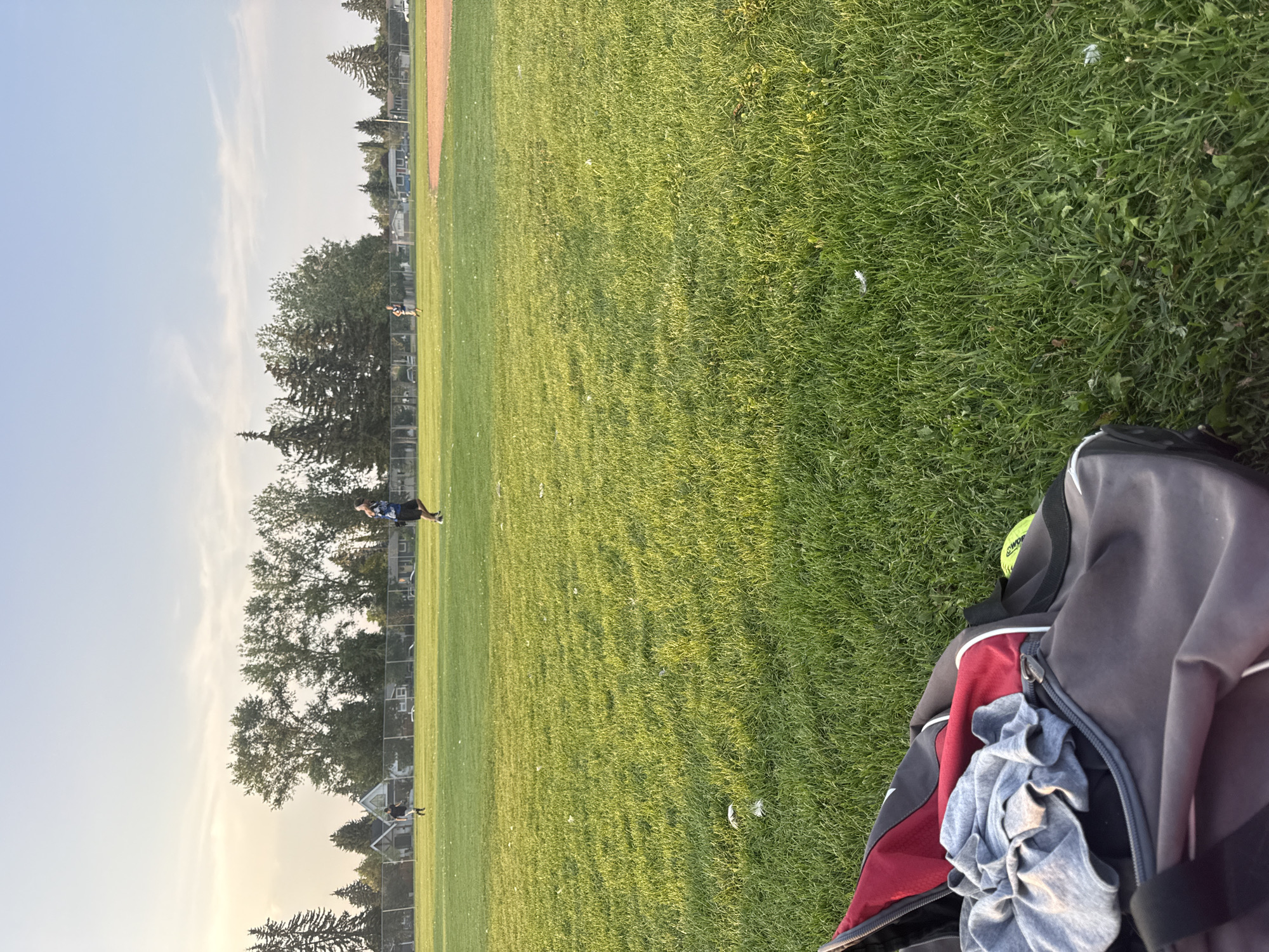 In Calgary, Canada, on a grassy field with a clear sky, two individuals are engaged in a game. One person is in the middle of a run, while another is standing at a distance. In the foreground, a duffel bag is positioned on the grass, with clothing and a yellow ball visible inside. The field appears to be part of a sports complex or park.