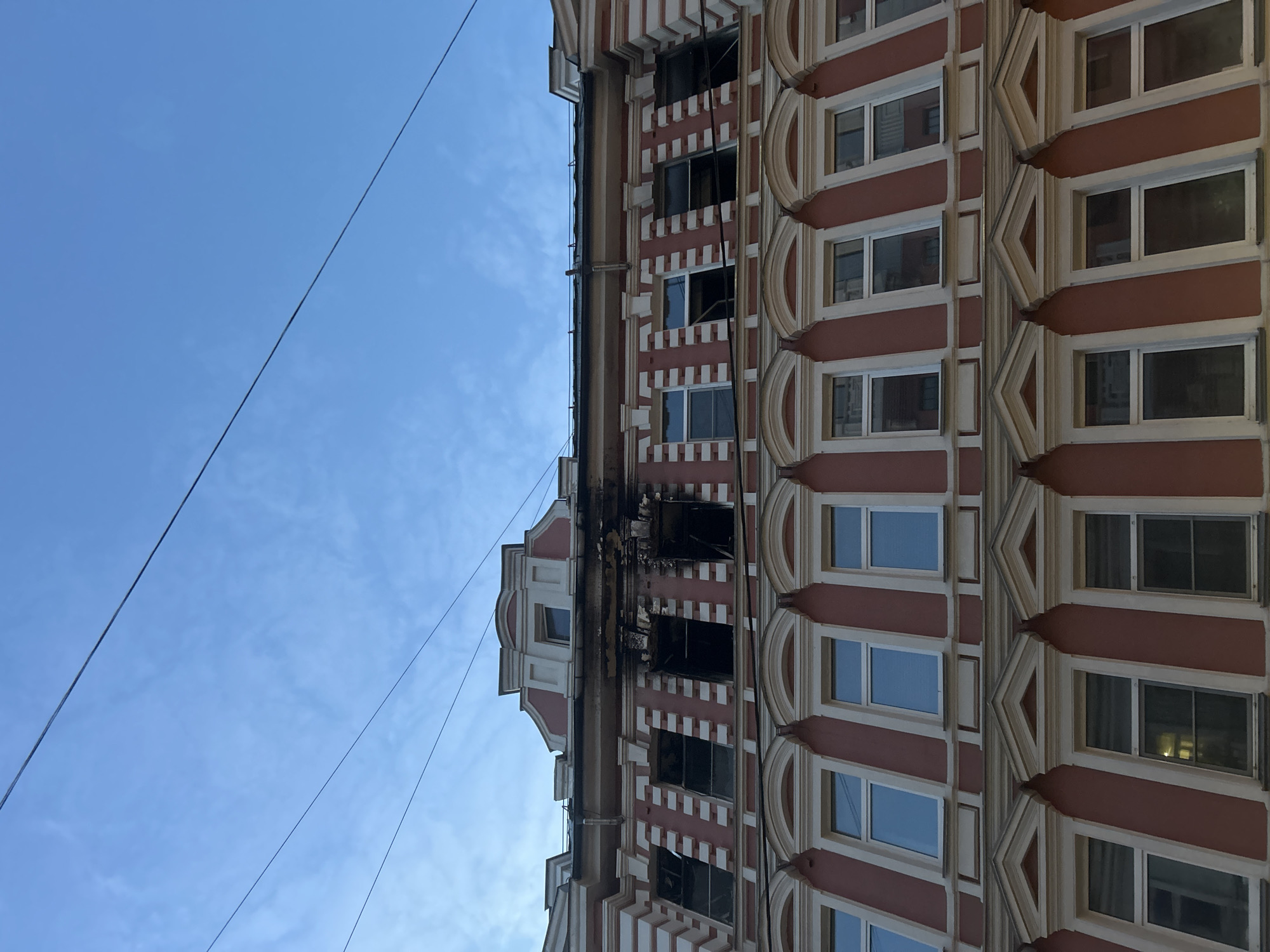 The image captures a partial view of a multi-story building, heavily damaged by fire, against a clear blue sky. The building features an ornate architectural style, characterized by a reddish-brown facade accentuated with white decorative elements, including vertical pilasters and textured patterns between windows, and curved moldings around some window frames.
A prominent section of the building, concentrated on the upper floors, shows extensive fire damage. Black soot covers large areas of the facade, particularly around several windows that appear completely burned out, shattered, or missing their glass. The charred marks extend vertically up the building from the affected windows, indicating the path of the fire.
The sky is a bright, clear blue with minimal, wispy cloud cover, suggesting daytime. Several thin, dark utility lines or cables crisscross the upper portion of the frame against the sky. No people or other objects are visible, focusing the viewer's attention solely on the damaged structure. The architectural style and evident damage suggest a historical building in an urban setting, located in Moscow, Russia.