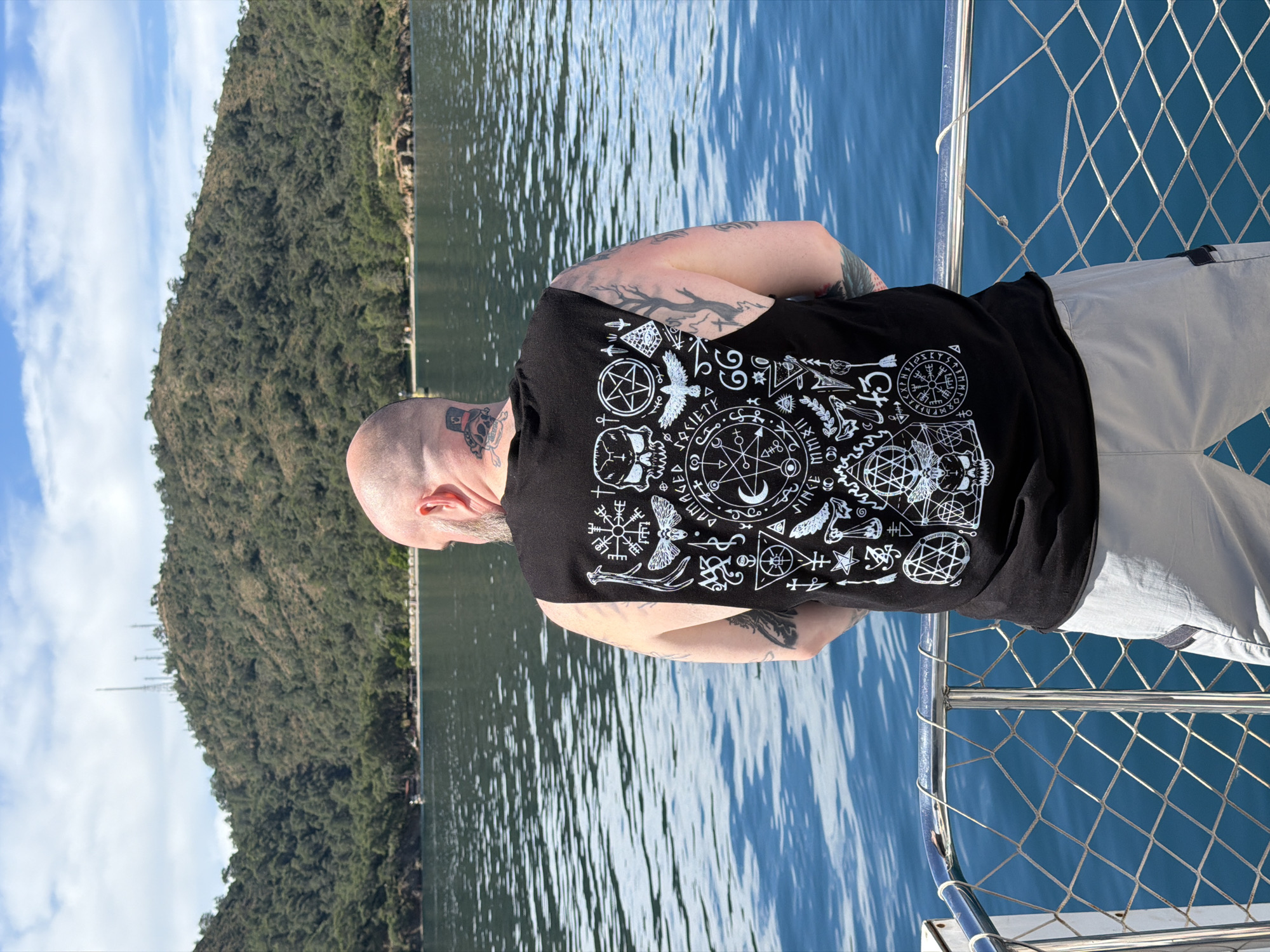 A man with tattoos is standing on a boat. He is wearing a black tank top with white occult-themed graphics, and light-colored pants. The boat is on the water, and a hillside covered in trees is visible in the background. The location is likely in the city of Fethiye, Turkey.
