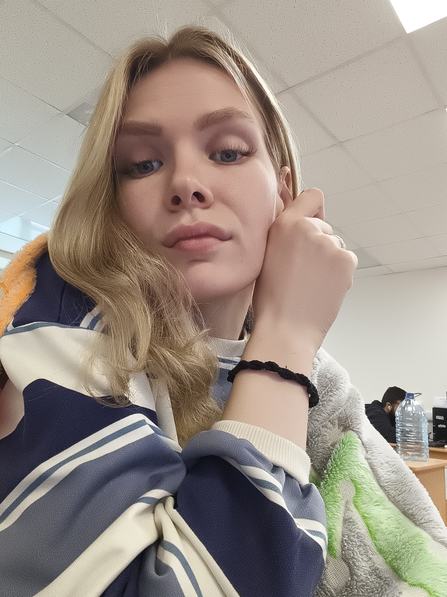 A medium-angle indoor shot presents a fair-skinned blonde woman with her face angled slightly downward. Her left hand is raised to her face, fingers touching her ear. A dark bracelet encircles her wrist. She wears a horizontally striped blue-and-white long-sleeved shirt with a plush, multi-colored shawl draped over her shoulders.
In the blurred background, a person sits at a desk, partially obscured. A large water bottle and office supplies are visible on the desk. The location is Izhevsk, Russia. The setting is an office or study environment with a drop ceiling.