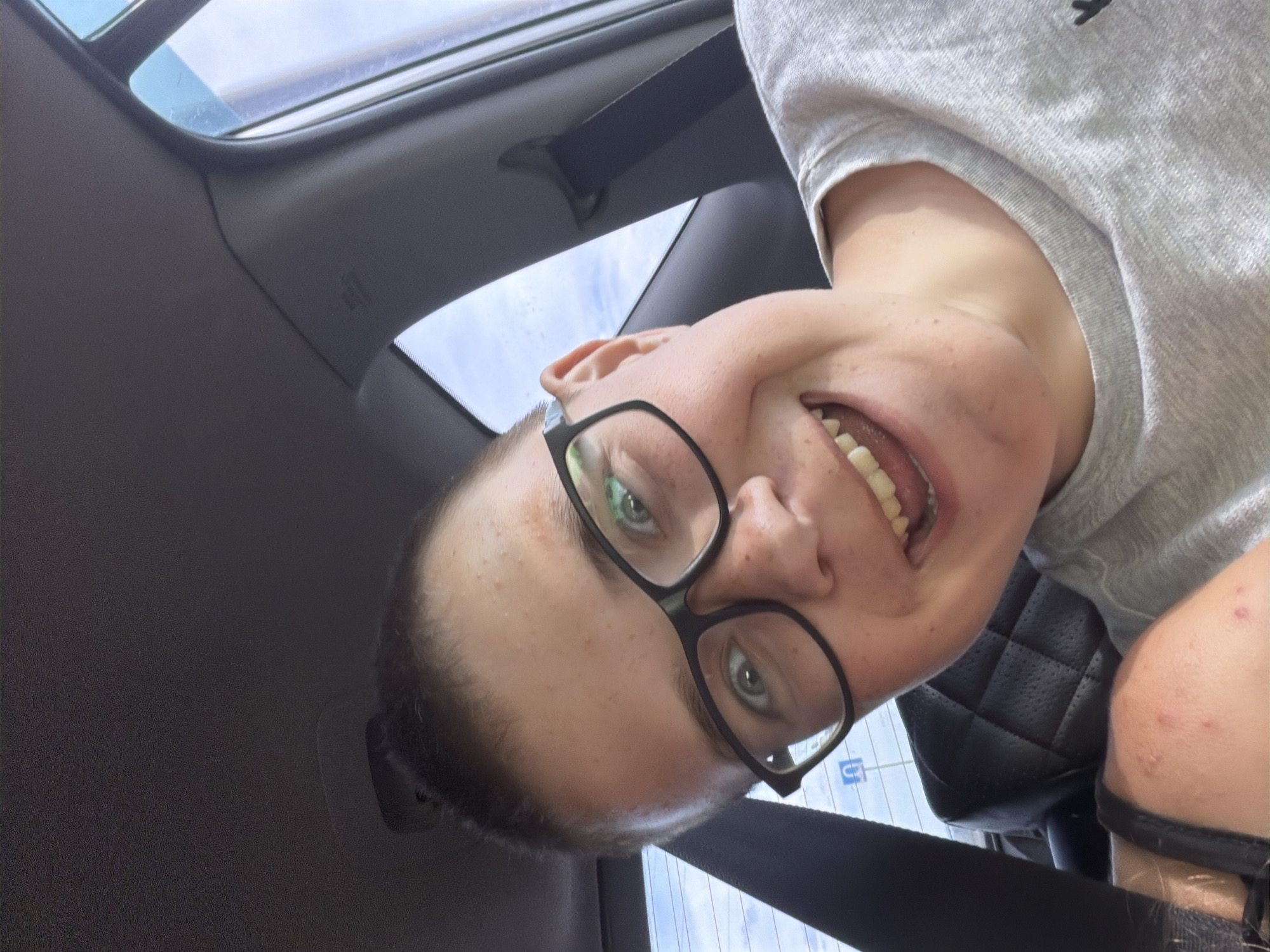 A person with fair skin, freckles, and light brown hair pulled back into a bun is taking a selfie in a car. They are wearing black-rimmed glasses and a light gray t-shirt. Their mouth is open in a wide smile, revealing their teeth. The background shows the interior of the car, including a black seat with a diamond pattern, a seatbelt, and the car's headliner. Outside the car window, a blue sky with white clouds is visible. The image appears to have been taken during the daytime. The location is noted as Novoye Mavrino, Russia, however, there are no visual cues within the image to confirm this specific location.