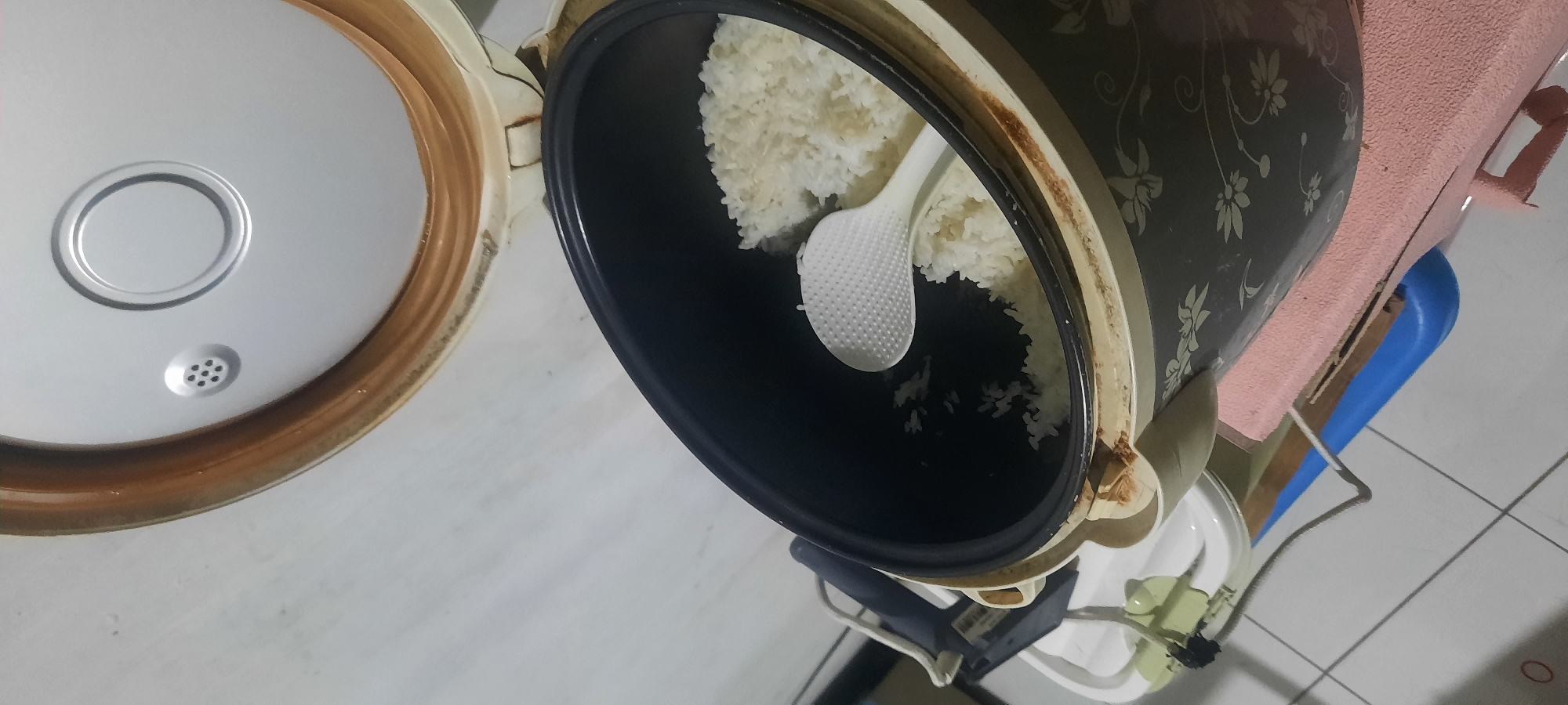 A rice cooker is open, revealing cooked white rice and a white rice paddle resting inside. The inner pot of the rice cooker has a black non-stick coating with some slight wear. The lid of the rice cooker is up, showing a silver interior with a circular indentation and a small grate. The exterior of the rice cooker is beige and decorated with a floral pattern. The rice cooker is placed on a pink textured mat, which is on top of a blue object and a wooden surface. The background appears to be a wall with a tiled floor. The image is oriented vertically.
