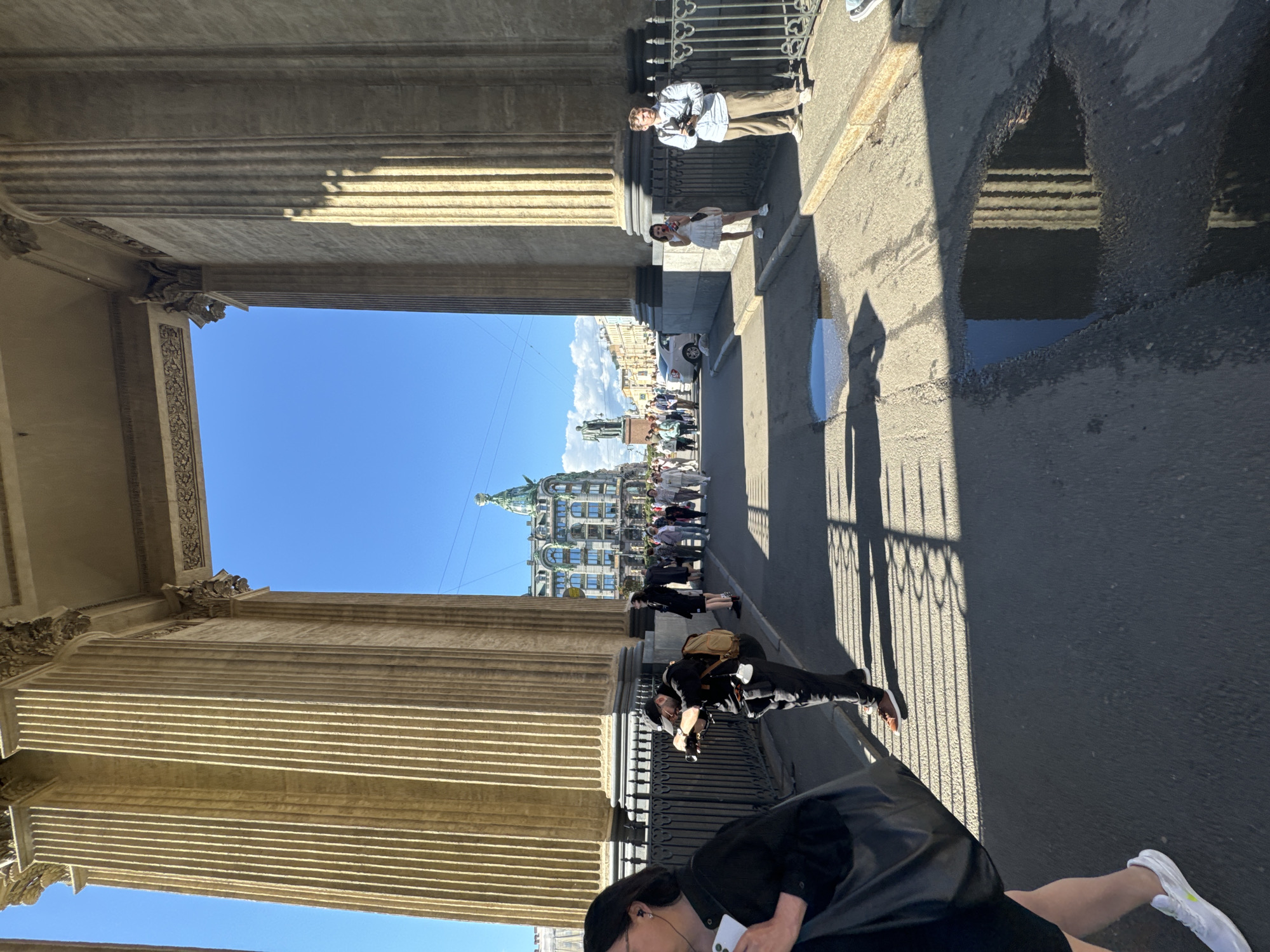 The image depicts a view through the colonnade of Kazan Cathedral in Saint Petersburg, Russia, on a sunny day. In the foreground, on the left, a woman in a black top and skirt is seen from the waist down, walking to the left, carrying a black bag. To her right, a man wearing a black t-shirt, black cap, and beige backpack is focused on taking a photo with a camera. Further down the walkway, a man in a light blue shirt and beige pants stands to the right, holding a camera. A woman in a white dress is to his left, holding a phone. Beyond the colonnade, a street is visible with pedestrians walking and a white car. In the background, a grand building with a green dome, a statue, and other architectural structures can be seen. The ground in the foreground has several puddles reflecting the sky and the colonnade.