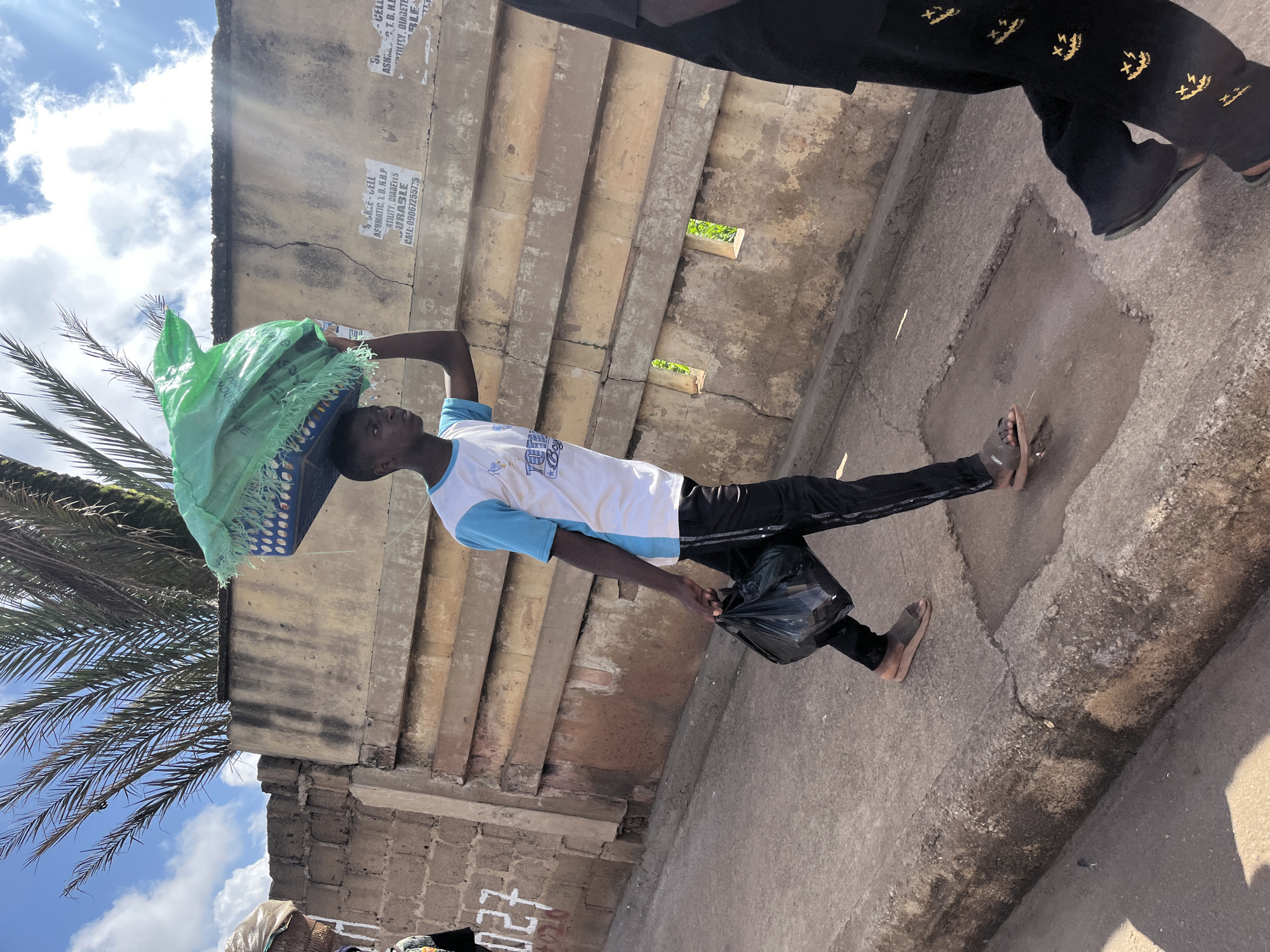 A young boy, appearing to be in his teens, walks along a dusty sidewalk in Jos, Nigeria, carrying a large green bag balanced on his head. He wears a white t-shirt with blue sleeves and black athletic pants adorned with a repeating pattern of yellow smiley faces. He also carries a black plastic bag in his right hand. The boy is walking past a weathered, light-colored concrete wall covered in graffiti and posters. Palm fronds are visible against a bright, cloudy sky. The environment suggests a bustling urban setting, possibly a market area given the boy's load and the surrounding wall, which may delineate property or offer shade. The overall scene captures a moment of daily life in the city.