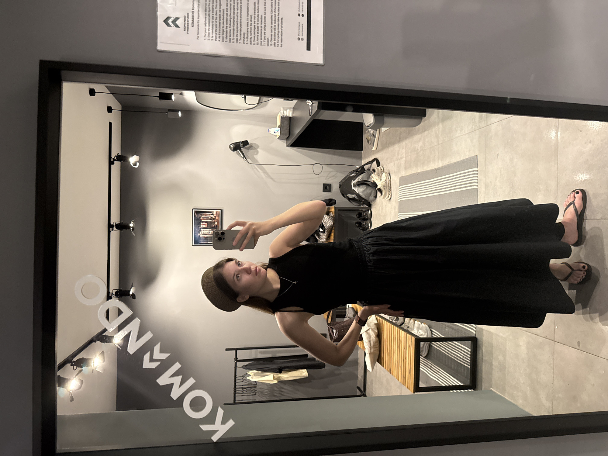 The image shows a woman taking a selfie in what appears to be a fitness studio. She is wearing a dark dress, a hat, and sandals. The studio, named "KOMANDO", has grey walls, a bench with storage, workout equipment, and signs, one of which is displayed in English. The text on the sign suggests this studio is located in Belgrade, Serbia. The photo is taken in a full-length mirror.