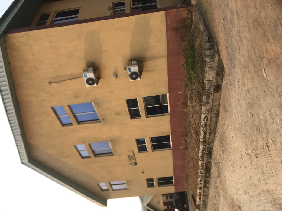 The image displays a multi-story building alongside an unpaved road, under what appears to be a sunny sky in Umudike, Nigeria. The building is predominantly light yellow or beige, featuring a dark red or brown trim along its lower facade and possibly under the roofline. A segment of a dark-colored, sloped roof with an overhang is visible on the left side.
Multiple rectangular windows with dark frames and reflective glass are visible across the building's walls. Two white outdoor air conditioner units are mounted on the upper part of the building, casting distinct shadows that suggest strong sunlight, indicating it is daytime, likely mid-morning or afternoon. Below a set of windows, a small circular logo or text, which appears to include the word "Bedford," is visible on the wall.
Adjacent to the building on the right is a rough, unpaved dirt road or path, light brown in color, with textures suggesting foot or vehicle traffic. A low, irregular stone or concrete curb separates the building's foundation from this pathway. Along the curb and the immediate ground next to the building, there is sparse, dry grass interspersed with some patches of green vegetation. The scene is static, with no people or specific activities taking place.
