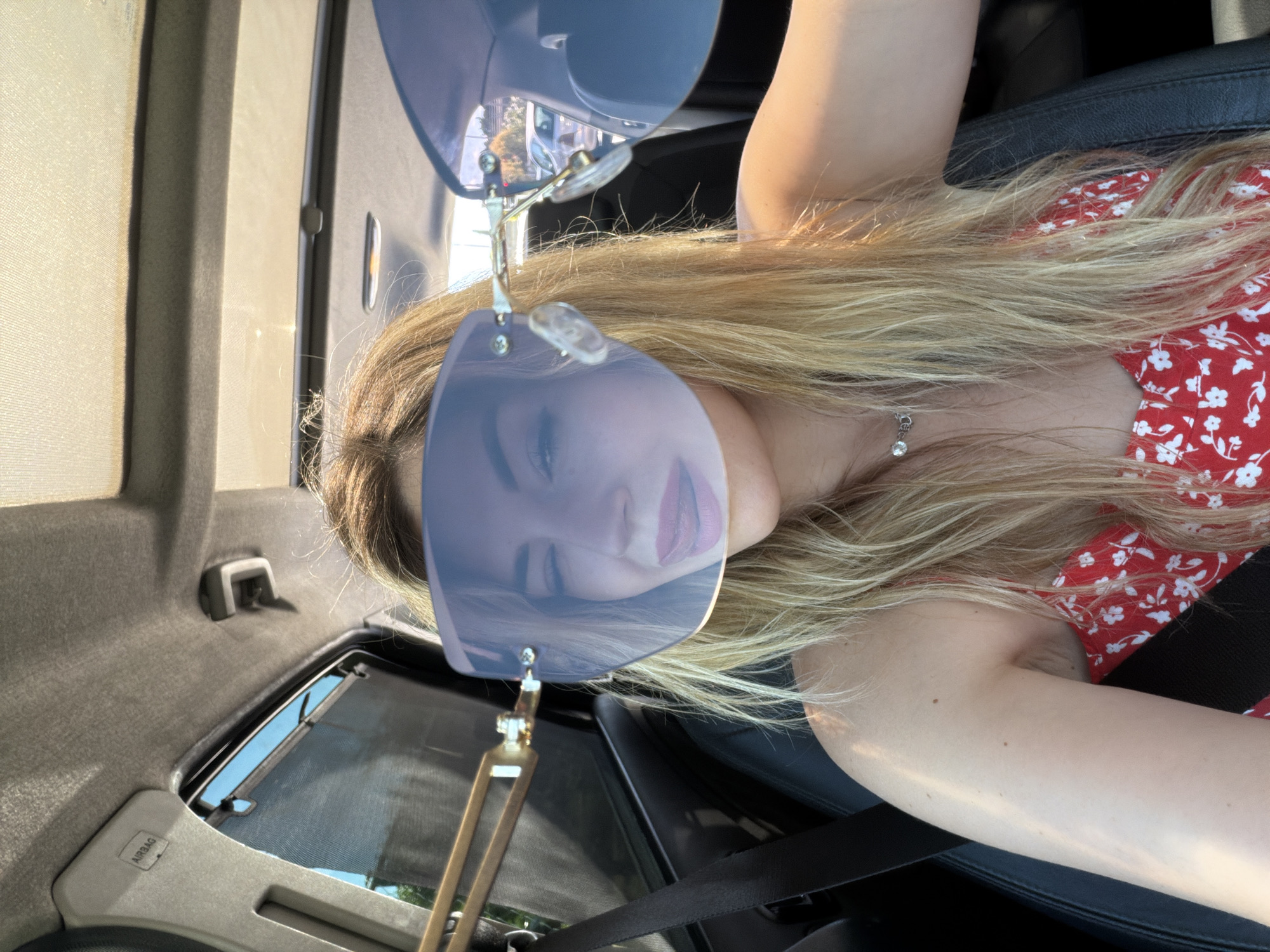 A young woman with long, wavy blonde hair is pictured inside a car in Podgorica, Montenegro. She is wearing a red top or dress adorned with a white floral pattern. Her face is largely obscured by large, blue-tinted mirrored sunglasses, which reflect a hint of her pinkish lips. A delicate silver necklace with a small, clear gemstone hangs around her neck. She appears to be reclining or lying down in the vehicle, with a black seatbelt fastened across her.
Strong, bright sunlight illuminates her hair and exposed arms, suggesting a clear, sunny day. The car's interior features dark upholstery for the seats and door panels, contrasting with a lighter grey headliner. Through the vehicle's windows, blurred external elements such as trees and a glimpse of the sky are visible. A retractable sunshade or screen is present on the rear side window. On the interior trim near the rear window, the word "AIRBAG" is clearly embossed. The image is oriented sideways, with the woman's head towards the top left of the frame and her body extending diagonally, indicating the photo may have been taken from an overhead or side angle and then rotated. The scene captures a casual moment inside the vehicle.