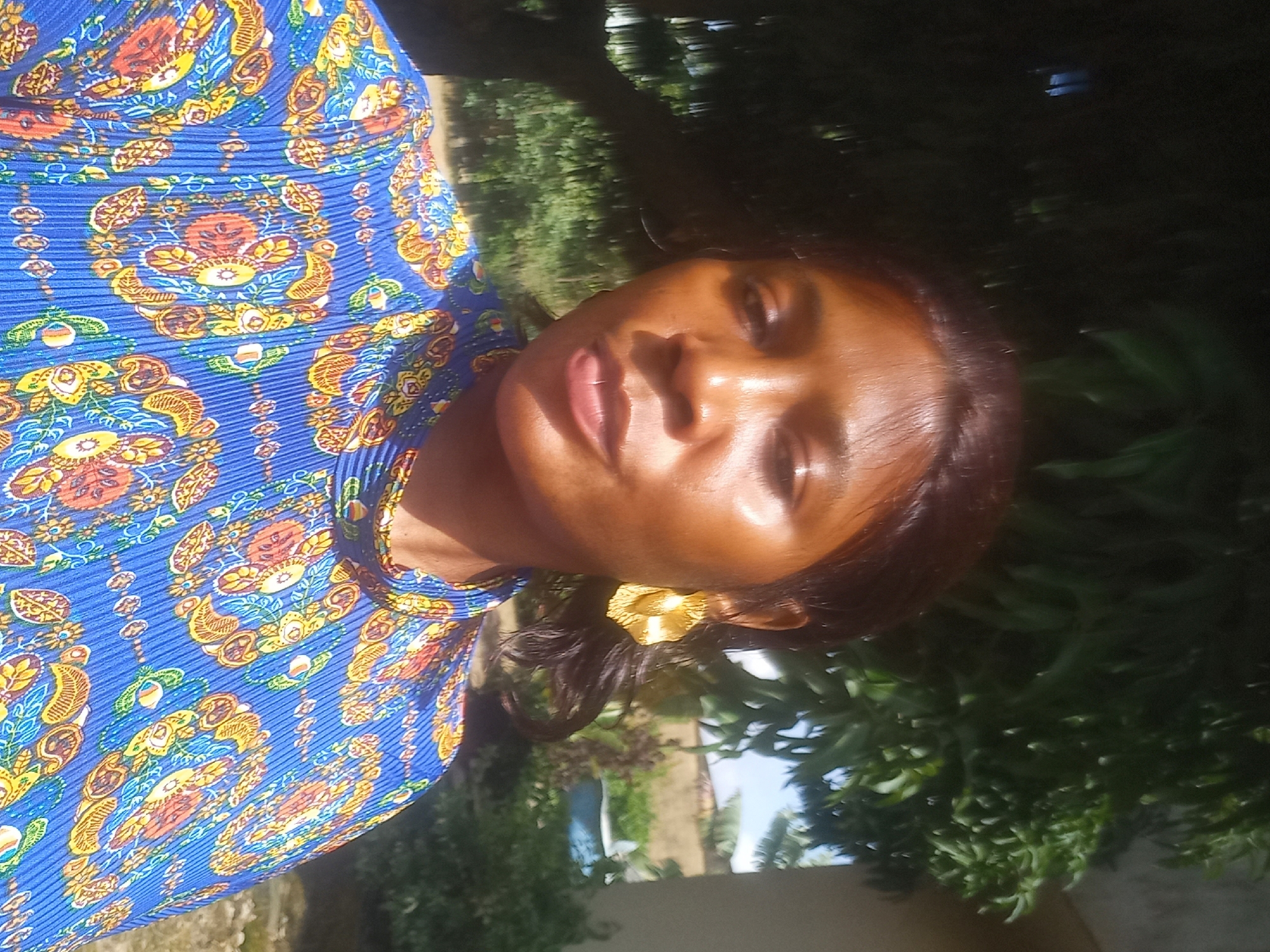 A medium shot shows a woman of African descent with dark hair and brown skin, wearing a blue shirt with a colorful, intricate pattern. She has large, gold earrings shaped like flowers. The woman is outdoors, with greenery in the background, including trees and bushes. The lighting suggests it is daytime. The woman's face is slightly tilted, and she appears to be looking at the camera.