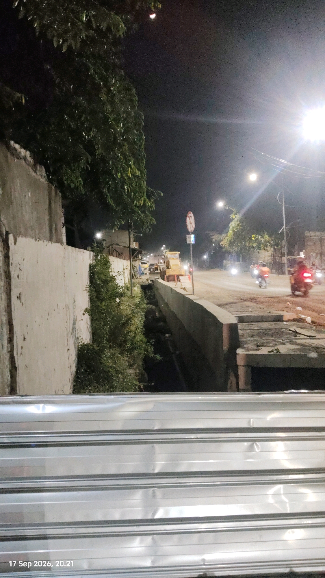 An outdoor, low-light photograph taken at night captures a street scene in Surabaya, Indonesia, potentially near a construction site. The foreground is dominated by a textured, metallic structure, possibly corrugated metal sheeting, which is slightly out of focus. A date and time stamp, "17 Sep 2026, 20.21", is visible in the bottom left corner, suggesting this is a recorded image from the future.
Beyond this metallic obstruction, the scene opens to a street. To the left, a tall, textured concrete wall runs parallel to the road, with dark foliage and trees growing alongside it. A shallow, dark trench or drainage channel runs along the base of the wall, bordered by a concrete structure.
Further down the street, construction equipment, including a yellow bulldozer, is visible. Streetlights illuminate the scene, casting a bright glow on the road and surrounding areas. Several motorcycles are in motion on the road, their headlights and taillights creating streaks of light. The road surface appears to be rough, suggesting ongoing construction or repairs. The overall atmosphere is one of urban nighttime activity, with a blend of construction, traffic, and existing infrastructure.