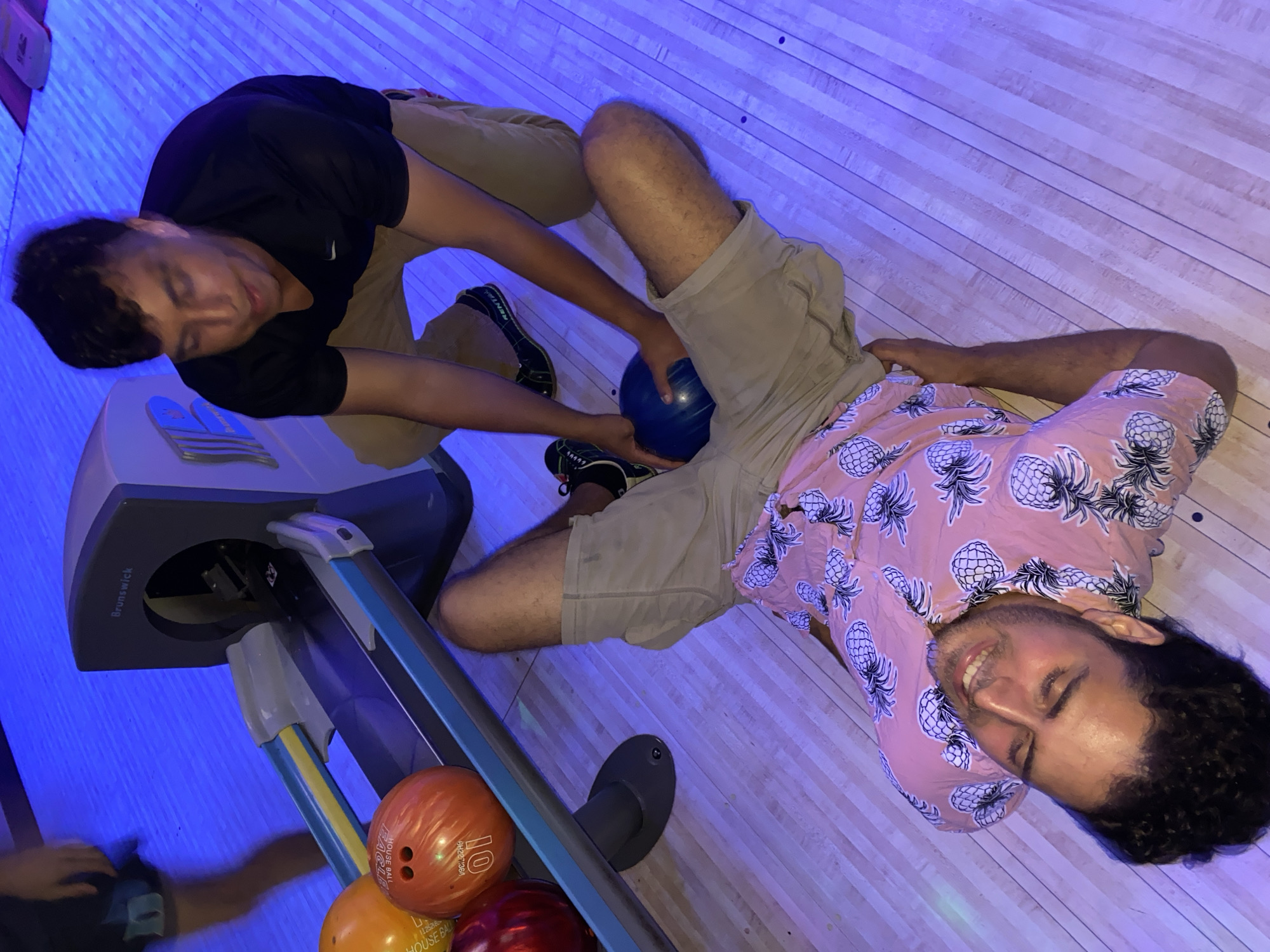 A person in a pink shirt with a pineapple pattern lies on their back on a bowling alley lane. Their legs are splayed and they are smiling with their eyes closed. Another person in a black shirt and tan shorts is kneeling next to them, holding a blue bowling ball. The bowling ball is positioned between the kneeling person's hands and the lying person's legs. In the background, a Brunswick bowling ball return machine is visible, with orange and red bowling balls in the rack. The bowling alley is lit with blue lights. The location is the city of Waynesboro, United States of America.