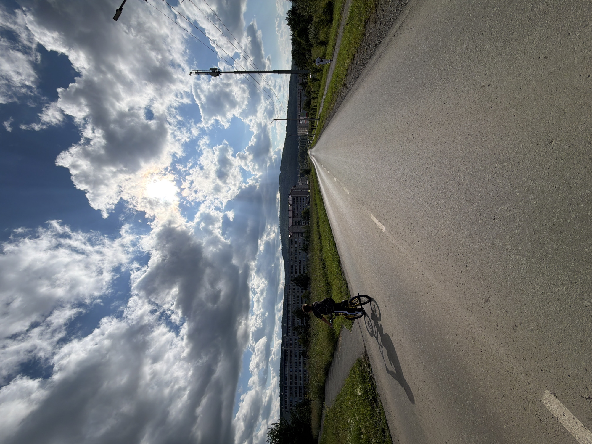 A low-angle, wide shot captures a paved road stretching into the distance under a dramatic sky filled with broken clouds. Sunlight pierces through the clouds, creating visible rays. On the left side of the road, a child on a bicycle casts a long shadow on the asphalt. The child is wearing a dark t-shirt and shorts, and the bicycle is yellow and black. Beyond the road and the child, a grassy field extends to a line of tall, grey apartment buildings. Trees are visible in the foreground and background. On the right side of the road, a narrow sidewalk runs parallel to the asphalt, with more grass and trees beyond it. A person in a white shirt and dark shorts walks away from the camera on the sidewalk. A utility pole with antennas and power lines stands on the right side of the road, with another pole and street light further down the road. The landscape beyond the buildings appears hilly and covered in trees.