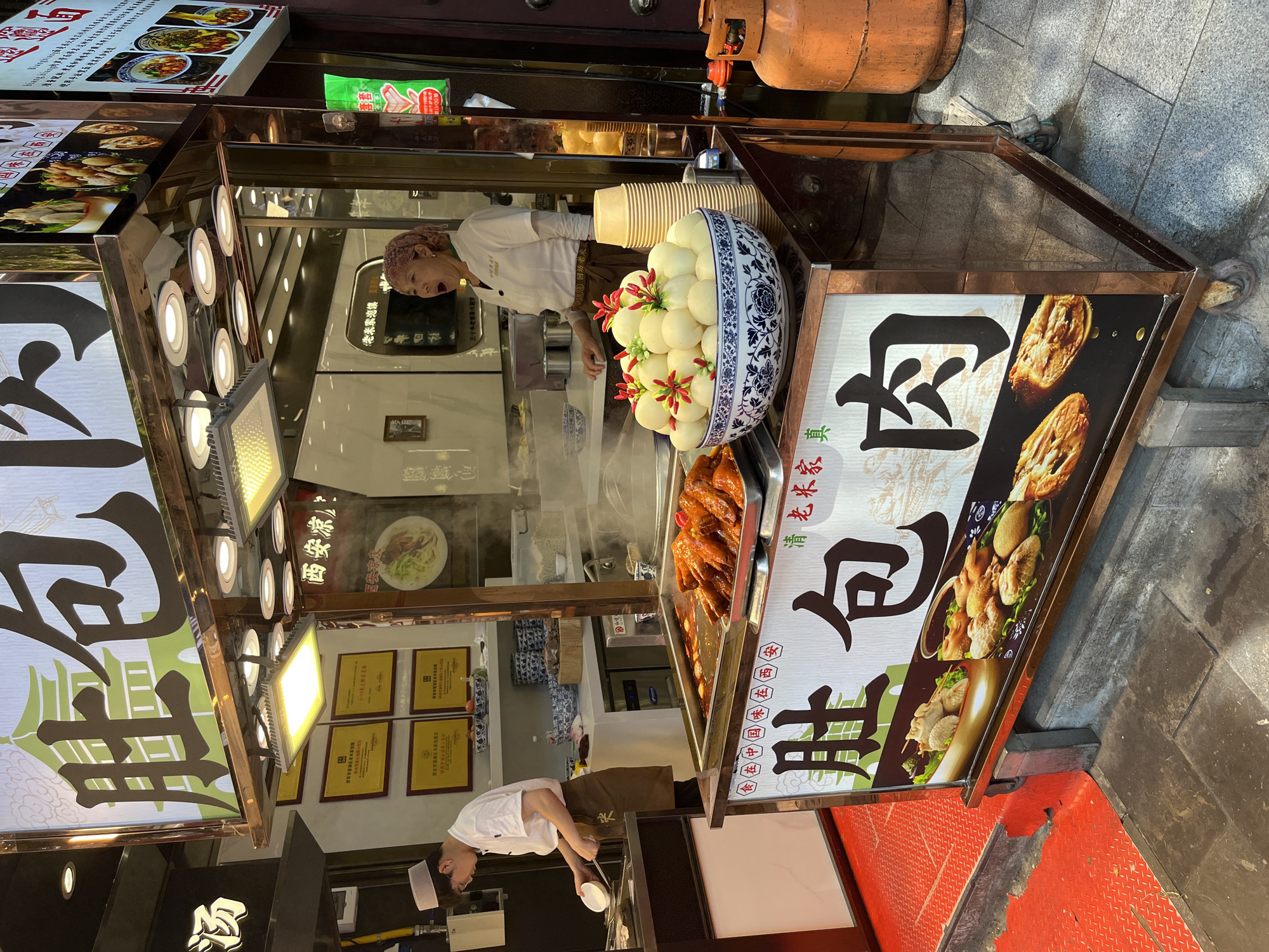 A street vendor stall in an urban environment sells steamed buns. A person wearing a white chef's coat and a brown apron stands behind a counter, preparing food. A second person, a woman wearing a white shirt and a pink headscarf, is visible inside the stall, yawning. In front of the stall, a large bowl overflows with white steamed buns, each topped with a small red and green garnish. A metal tray filled with glistening, cooked chicken wings is situated next to the bowl. A large sign above the stall displays Chinese characters and images of food. A propane tank sits to the right of the stall. The ground in front of the stall is a mix of red metal grating and grey cobblestones. The background reveals more of the urban setting, with a storefront visible to the right.