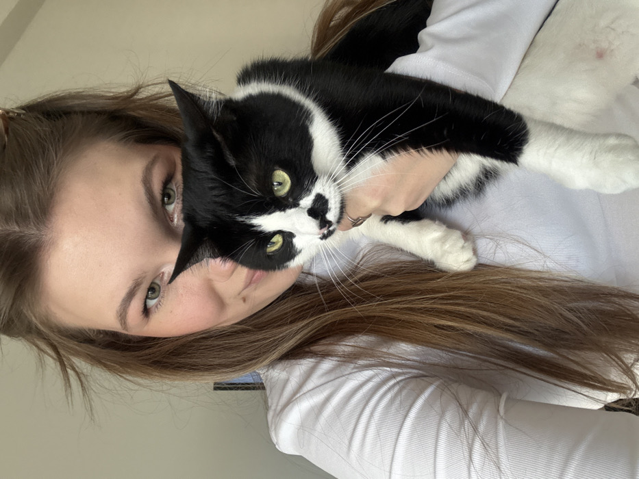A portrait-oriented image shows a young adult female holding a black and white tuxedo cat in an indoor setting in Belgrade, Serbia. The female, positioned on the left and upper frame, has long, light brown hair, green eyes, and a faint smile, looking directly at the camera. She is wearing a white long-sleeved top. Her left hand, visible and wearing a ring on the index finger, supports the cat. The cat, occupying the right and lower frame, features a black coat with white chest, belly, paws, and a white blaze on its face, notably including a distinctive irregular black patch on its nose and upper lip. The cat has green eyes and also looks towards the camera. The background is a plain, light-colored wall, indicating an indeterminate indoor environment.