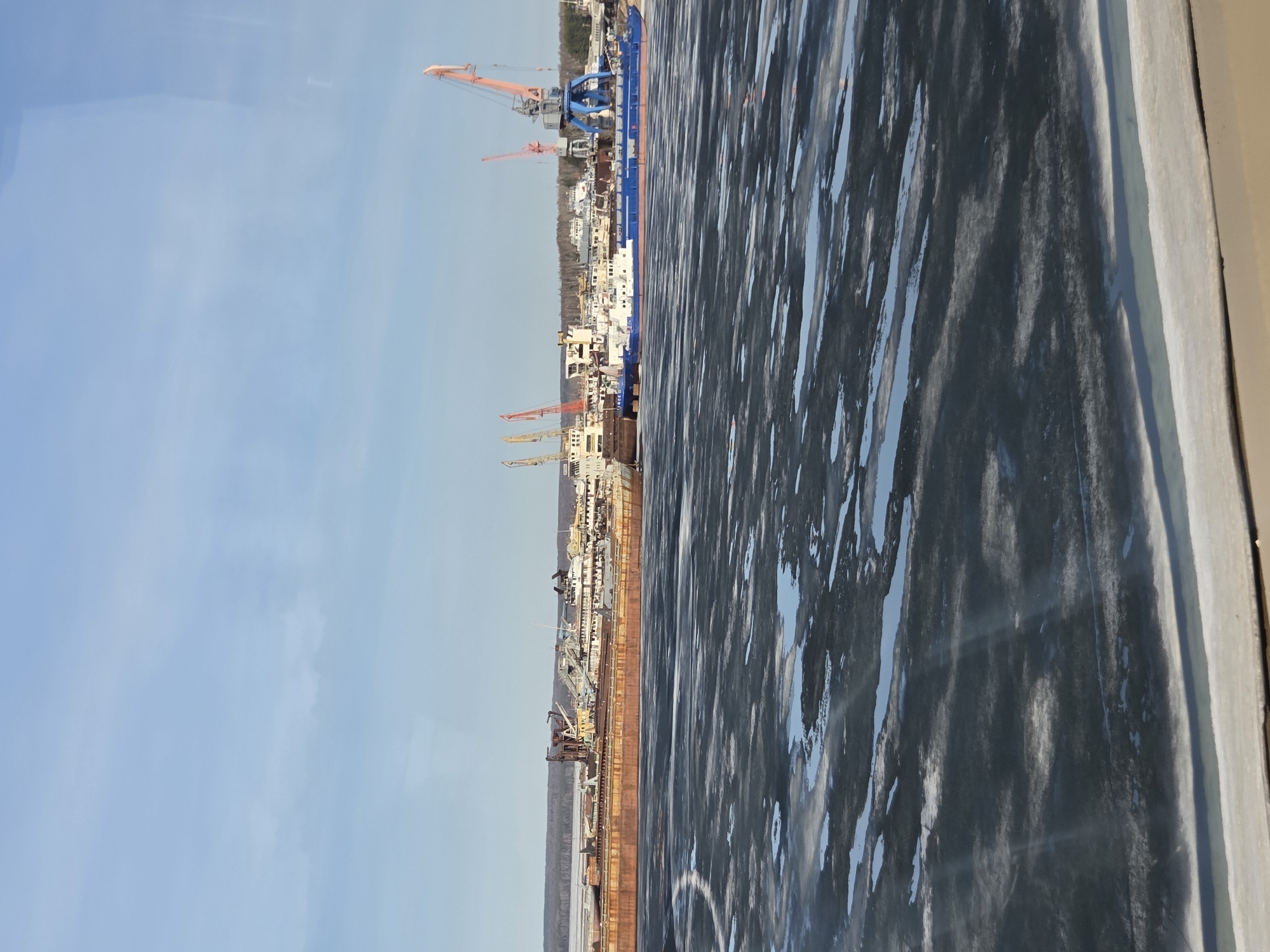 The image depicts a cityscape with a partially frozen body of water in the foreground, possibly the Kama River, consistent with the city of Chaikovsky, Russia. Several industrial cranes are visible along the waterfront, indicating a port or shipyard area. Buildings of varying heights are situated behind the cranes. The sky is mostly clear with a light blue hue. The frozen surface of the water is textured with ice and patches of open water.
