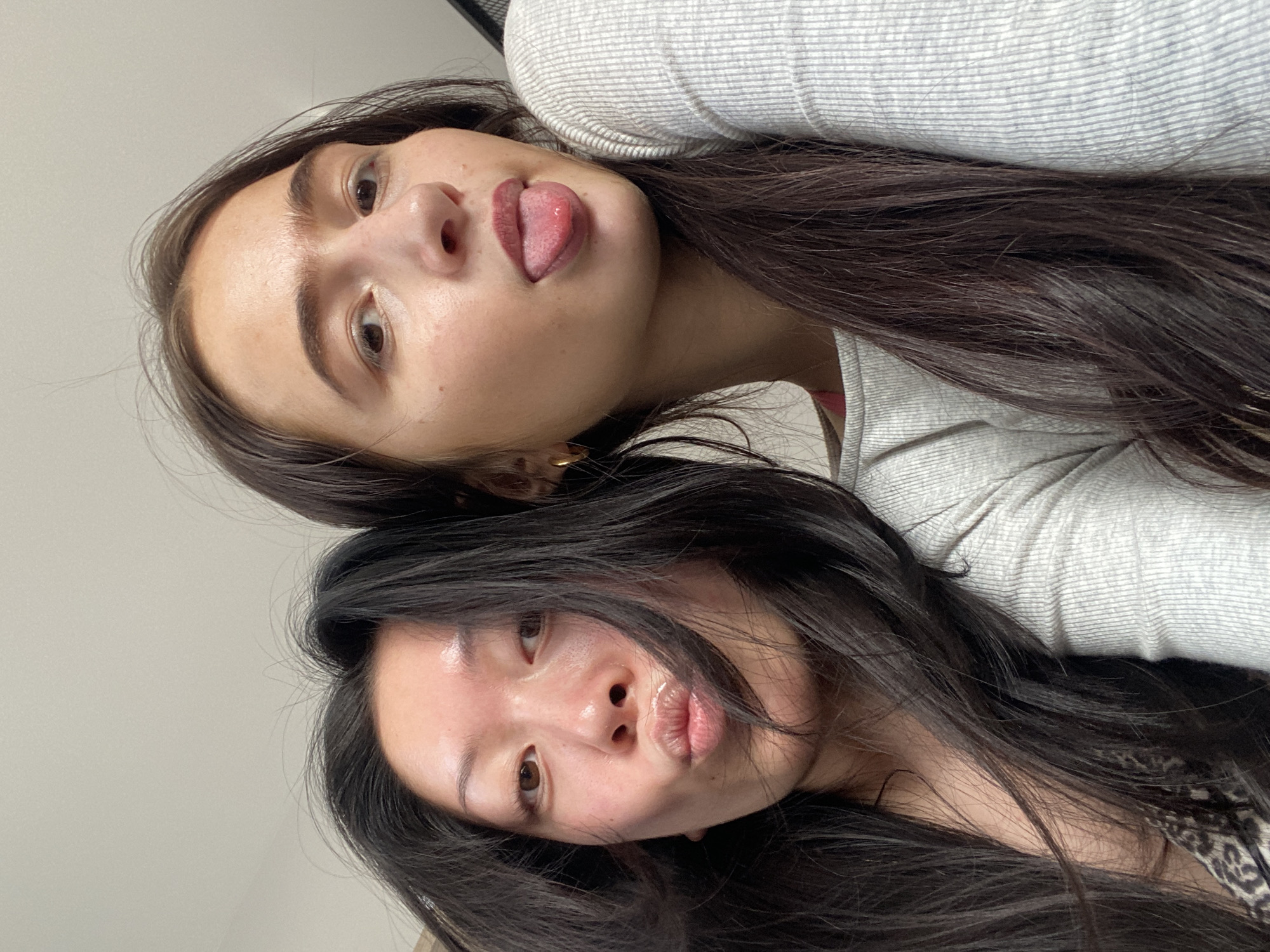 Two women are posing closely together for a photograph, taken at a low angle. The woman on top has dark hair and is sticking her tongue out. She is wearing a gray striped top. The woman on the bottom has dark hair and is pouting. The background is a light gray wall. The location appears to be indoors. There is no indication that the location is in London, United Kingdom.