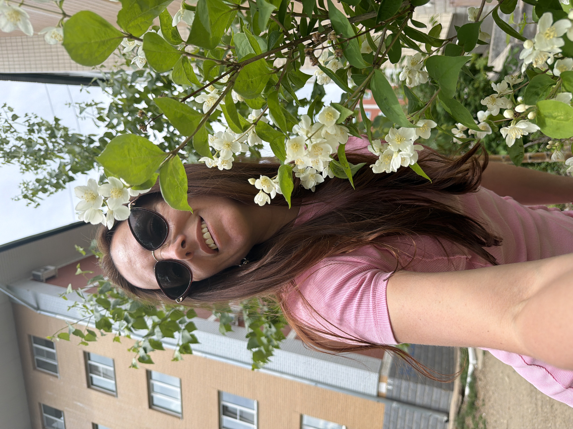 A young woman is taking a selfie outdoors. She is wearing a pink, short-sleeved, ribbed t-shirt and dark sunglasses. Her long, dark brown hair is down, and she is smiling at the camera. She is positioned in front of a blooming jasmine bush with white flowers and green leaves. In the background, there is a light brown apartment building with several windows and a grey and white section of another building. The ground in the foreground is dirt and some patchy grass. The scene appears to be in an urban courtyard in Birobidzhan, Russia.