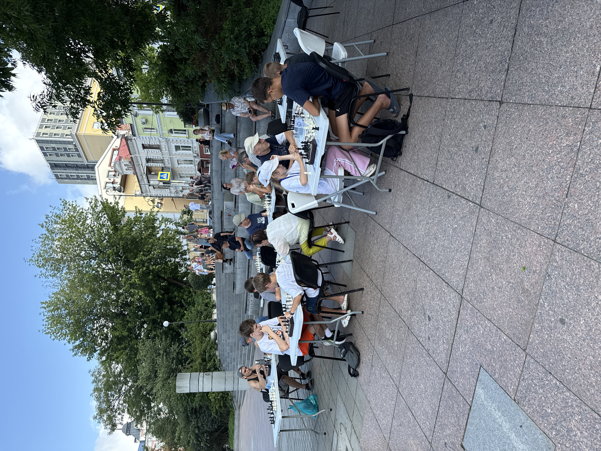 The image depicts an outdoor chess tournament or gathering in a public space. Several individuals, including adults and children, are seated at tables playing chess. White folding tables are set up, and chessboards with pieces are arranged on them. The participants are engaged in their games, with some looking intently at the board, while others appear to be in conversation. The setting appears to be a paved plaza with steps leading up to a higher level, suggesting a park or a public square. In the background, trees and buildings are visible, indicating an urban environment. One building in the background appears to be a modern high-rise, contrasting with older, more colorful structures. The overall atmosphere suggests a leisurely and engaging community event. This scene is located on the Triumphal Arch Hill in Vladivostok, Russia, known for its panoramic views and public spaces that often host cultural and recreational activities.