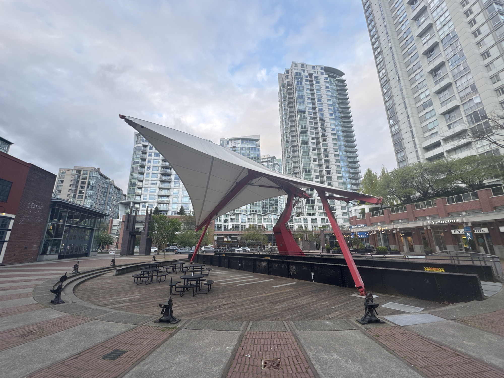 This image depicts a modern urban plaza in Vancouver, Canada, characterized by a striking architectural tensile structure. The central feature is a large, pointed white canopy, held aloft by prominent red support beams, creating a focal point over a circular wooden deck. This deck is furnished with several black picnic tables and benches, suggesting a public gathering or dining area. Decorative cast-iron bollards or anchors are spaced around the perimeter of the deck.
In the background, several tall, contemporary high-rise buildings with many windows dominate the skyline, reflecting the cloudy, overcast sky. To the immediate left, a brick building with large glass windows is visible. To the right, a multi-story brick building complex houses various businesses, with "FYIdoctors" and "coast to coastal" clearly visible on their storefront signs. Another sign partially shows "FYI S A". Scattered trees and greenery are interspersed among the buildings and around the plaza. The sky is uniformly cloudy, suggesting an overcast day with soft, diffuse lighting. The scene appears quiet and orderly, showcasing a functional public space within a dense urban environment.