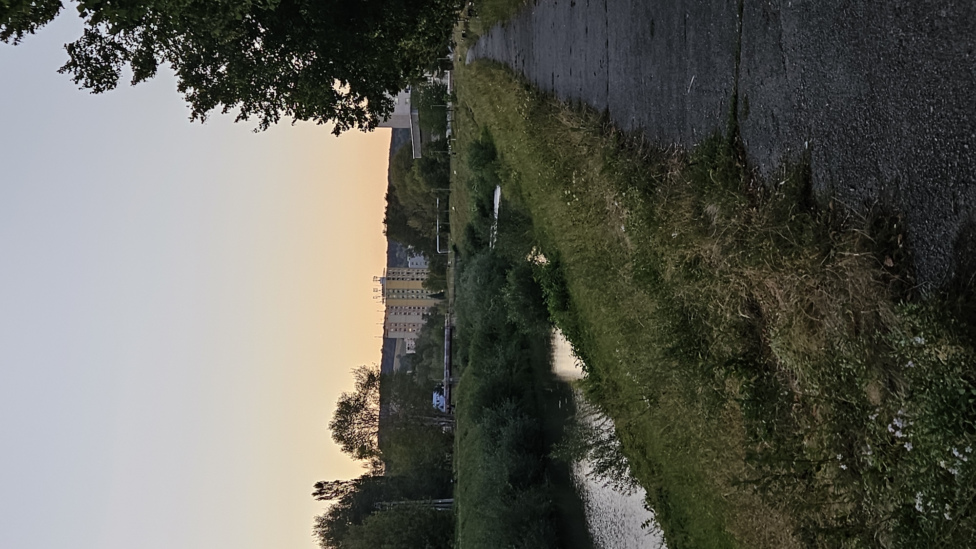 The image shows a paved walkway running alongside a river in the city of Prešov, Slovakia. The walkway is on the right side of the image, and the river is on the left. The riverbanks are covered in lush green grass and vegetation. In the background, there are several apartment buildings, some of which are tall and appear to be of Soviet-era architecture, and a hill. The sky is a pale orange and blue, indicating that it is either sunrise or sunset. There are no people or animals visible in the image. The scene is peaceful and serene. The image appears to be taken from the Sibírska neighborhood of Prešov, given the proximity of the river and the characteristic apartment buildings.