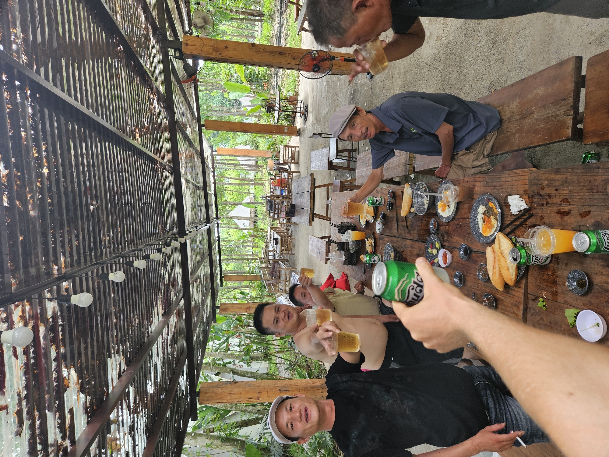 In Da Nang, Vietnam, a group of men are gathered around a long wooden table, seemingly enjoying a meal and drinks under a corrugated metal roof. The setting appears to be an outdoor, rustic eatery, possibly in a rural or semi-rural area given the surrounding lush greenery and trees. The roof, made of corrugated metal sheets, is supported by wooden pillars. Several bare light bulbs are strung along the underside of the roof, suggesting it might be evening or that the area is dimly lit.
The men are diverse in age and attire, all appearing to be of Asian descent. Some are holding glasses of beer or other beverages, and some are raising them in a toast, indicating a convivial atmosphere. Food is visible on the table, including bread, plates with what looks like eggs and other accompaniments, and several cans of beer, one prominently in the foreground. Two of the men are shirtless, while others are wearing casual shirts and caps.
In the background, more tables and chairs are set up, and the environment suggests a natural setting with abundant vegetation and trees. There are also glimpses of some white tent-like structures in the distance, which could be part of the establishment or nearby accommodations. The ground appears to be a mixture of concrete and dirt. The overall scene captures a moment of relaxation and social gathering.