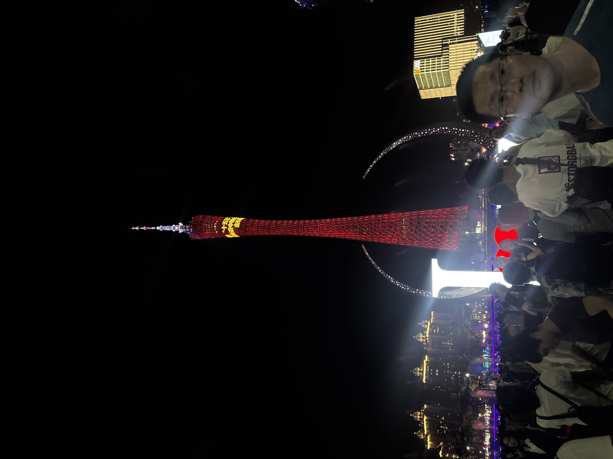 This image depicts a nighttime scene in Tianhe District, China, likely a public gathering or celebration. In the foreground, a crowd of people is visible, with some individuals looking towards the camera. One person in the upper right quadrant is clearly in focus, wearing glasses and a white t-shirt.
Dominating the background is a tall, illuminated tower, possibly a landmark, glowing with red and yellow lights. The tower's intricate lattice structure is discernible. In the middle ground, there are illuminated buildings with a distinctly modern architectural style, possibly skyscrapers, featuring a grid-like pattern of lights. A curved, illuminated structure, adorned with small white lights, arches across the scene, adding to the festive atmosphere. The overall impression is one of a vibrant urban environment at night, with crowds of people enjoying a festive event. The presence of a large tower and modern buildings suggests a significant cityscape. The atmosphere appears lively, with many individuals gathered, likely for a specific event or celebration. No specific weather conditions are evident, but the darkness indicates nighttime.