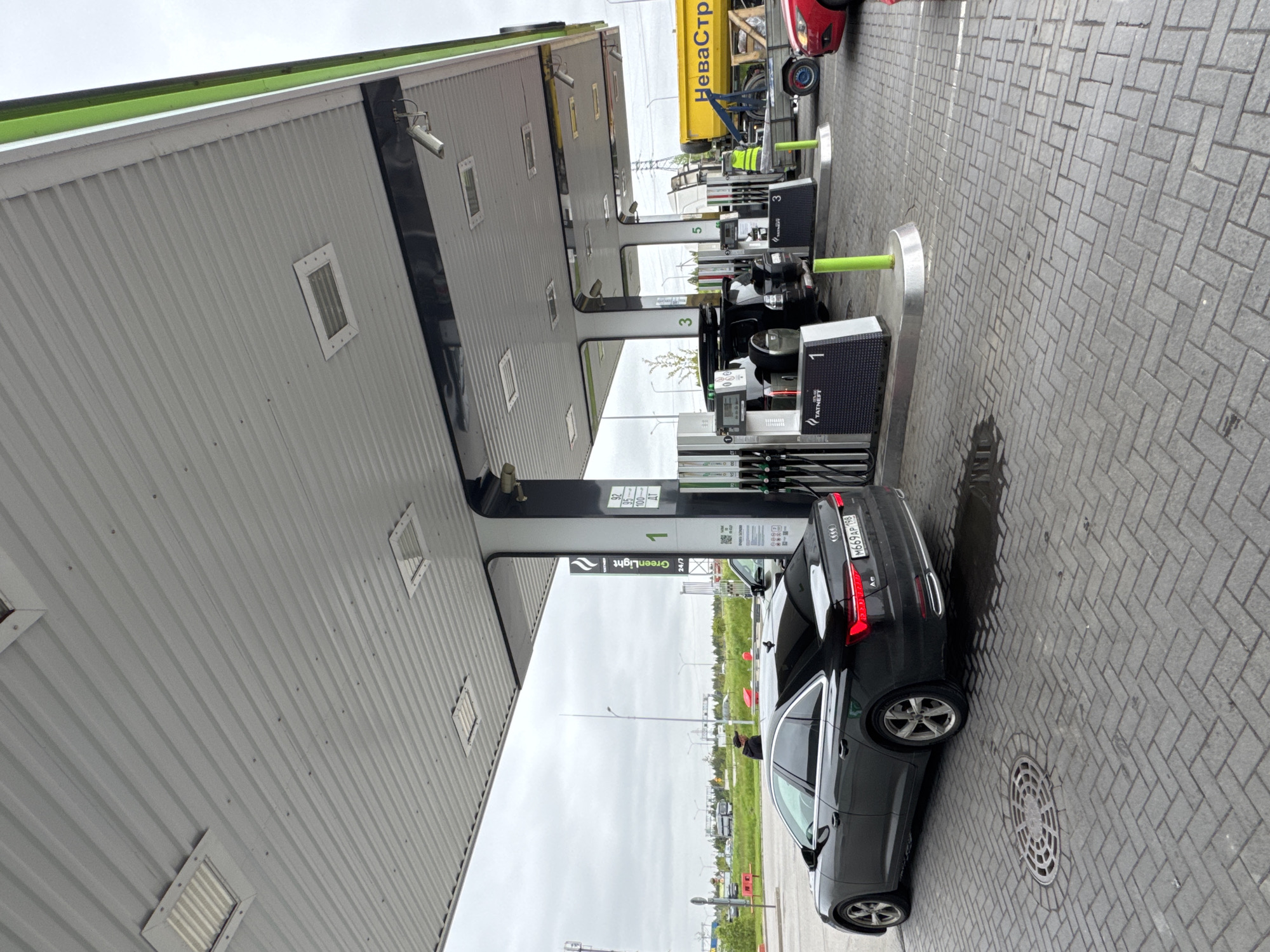 The image shows a "Green Light" gas station in what appears to be Medovoye, Russia. A black Audi sedan, with license plate number M669AP 198, is parked at pump number 1. Gas prices for 92, 95, and 100 octane fuel are listed on the pump. In the background, there are other pumps numbered 3 and 5, as well as a yellow truck with "НеваСтр" written on its side, possibly an equipment or maintenance vehicle. The weather appears overcast. The station building has a gray corrugated metal exterior with black accents and surveillance cameras. The foreground is paved with gray bricks.