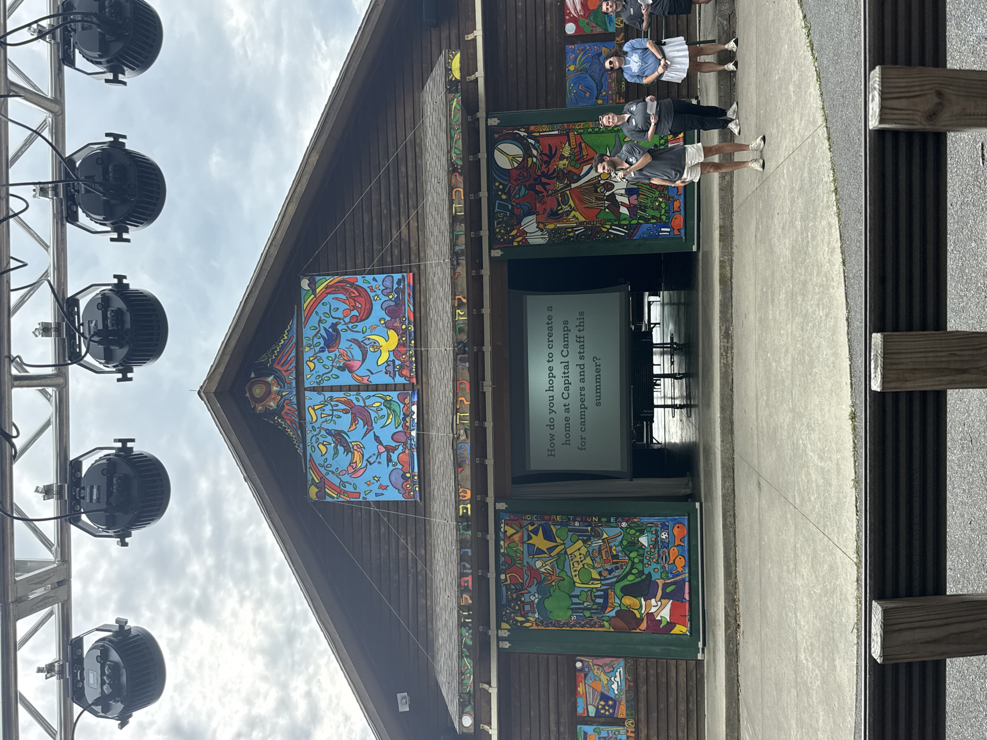 The image depicts an outdoor amphitheater stage, possibly at a summer camp, in Washington Township, United States of America. Above the stage, stage lighting fixtures are suspended from a metal truss. The backdrop of the stage is a wooden building with vibrant, colorful murals adorning its facade. A large screen is projected with the text, "How do you hope to create a home at Capital Camps for campers and staff this summer?". Several people are standing on the stage in front of the screen. One man in a grey polo shirt and white shorts is speaking into a microphone. To his right, a woman in a grey t-shirt and black pants holds a piece of paper. Further to the right, a woman in a light blue t-shirt and white skirt also holds a paper, and next to her, a man in a black shirt and pants stands holding a paper. The stage area is paved with concrete, and a small, grassy area is visible to the right of the stage. In the foreground, wooden posts and a metal barrier suggest a seating area for an audience. The sky is overcast with clouds.
