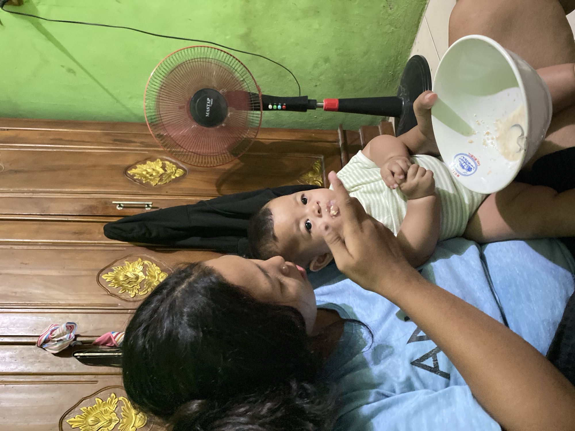 In a room in Tedunan, Indonesia, a woman is feeding a baby from a bowl. The woman has dark hair and is wearing a blue t-shirt. The baby is wearing a striped onesie. A fan is visible in the background, as is a wooden cabinet. The walls are painted green.