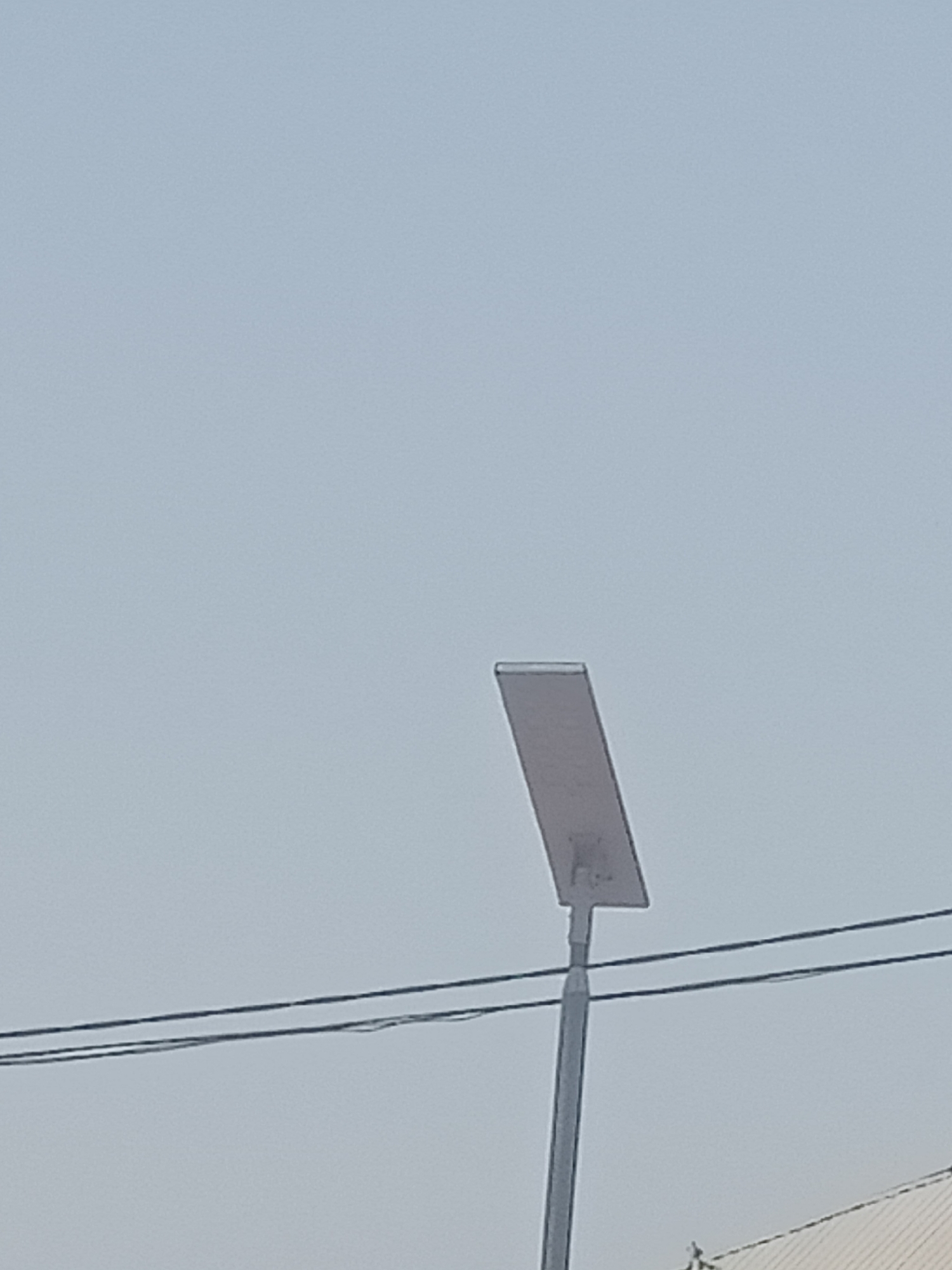 An outdoor, waist-up shot shows a solar-powered street lamp against a pale blue sky. The lamp is positioned to the right of the frame, with its panel angled towards the viewer. Two dark wires run horizontally across the mid-ground of the image. The lower right corner of the image shows a sliver of a white roof with a textured surface. The image is taken in Jalingo, Nigeria.