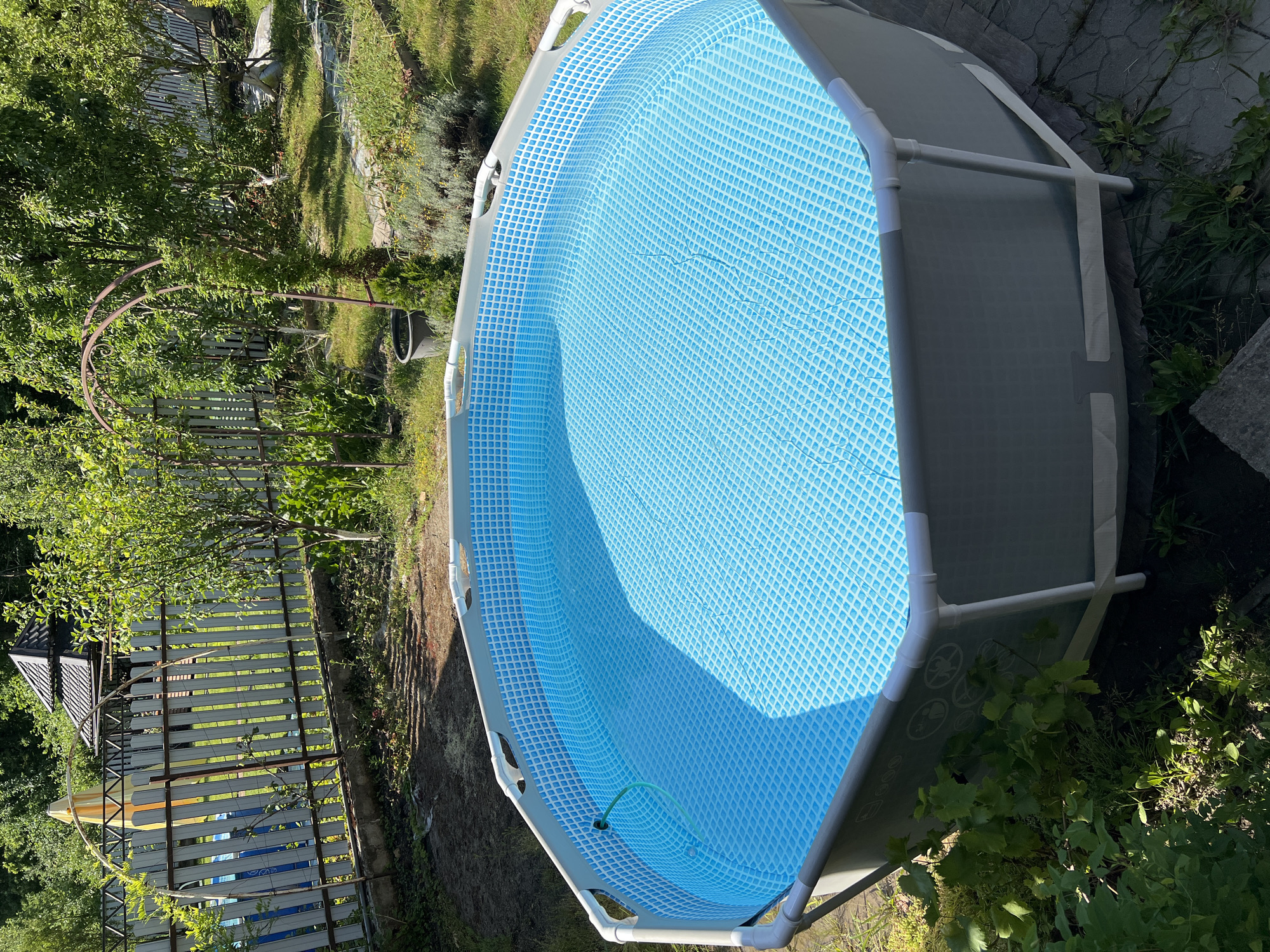 In an outdoor setting, likely a backyard in Goryachiy Klyuch, Russia, an octagonal above-ground swimming pool filled with clear blue water is the primary subject. The pool has a gray outer wall and white connectors. A green hose is visible entering the pool on the left side. The pool is situated on a paved surface, surrounded by grass, and a variety of green plants and trees. In the background, a white picket fence encloses a yard with a decorative gazebo or covered structure. Sunlight casts shadows on the ground and reflects off the water's surface, indicating a bright, sunny day. The scene is peaceful and suggests a leisure activity.