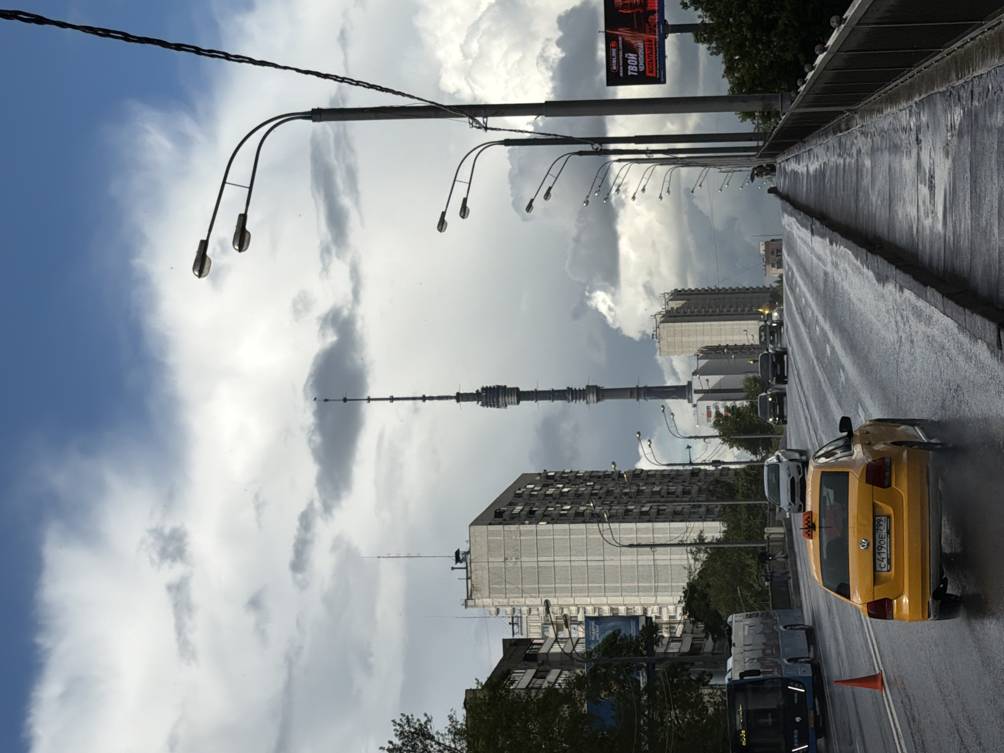 This media file captures a street scene on a cloudy day in Moscow, Russia. The perspective is from a moving vehicle, looking down a wet asphalt road lined with streetlights. In the foreground, a bright yellow taxi with visible license plates (C419OE 799) is a prominent object. Further down the road, other vehicles, including a bus, are visible, suggesting traffic. The background is dominated by a mix of urban architecture, including tall apartment buildings with numerous windows, and a striking television tower in the distance. The sky is dramatic, filled with large, billowing clouds that create a sense of depth and impending weather. The wet road surface indicates that it may have recently rained or is currently raining lightly. The overall mood is somewhat somber due to the cloudy conditions, but the vibrant yellow taxi adds a touch of color to the scene. The presence of the television tower and the Cyrillic text on the taxi's roof sign (likely indicating a taxi service) provide strong cues to the location being Moscow. The scene appears to be during daytime, given the illumination and visibility.