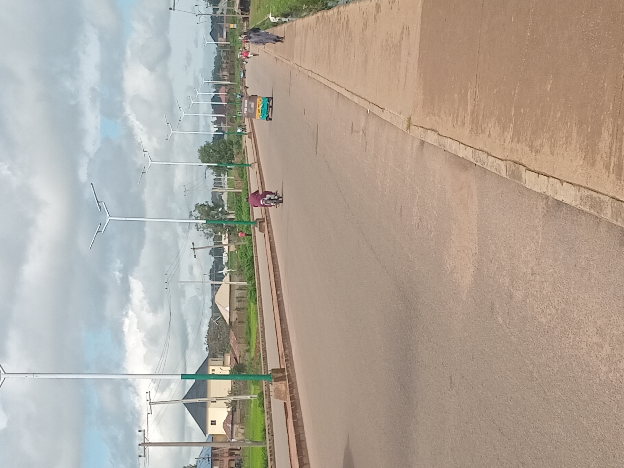 A wide, paved road runs through a somewhat suburban area, likely Bukuru, Nigeria, under a cloudy sky. The road is lined with modern streetlights on tall poles. On the left side of the road, there are residential buildings with distinct architectural styles, some with pitched roofs. A motorcycle carrying a single rider in a maroon outfit travels down the road. Further down, a colorful auto-rickshaw is also moving. On the right side of the road, a concrete sidewalk runs alongside, and a few individuals are walking. One person in a light-colored garment is walking towards the foreground, while others are further up the road. The environment appears to be a developing urban or semi-urban area with scattered vegetation and infrastructure.