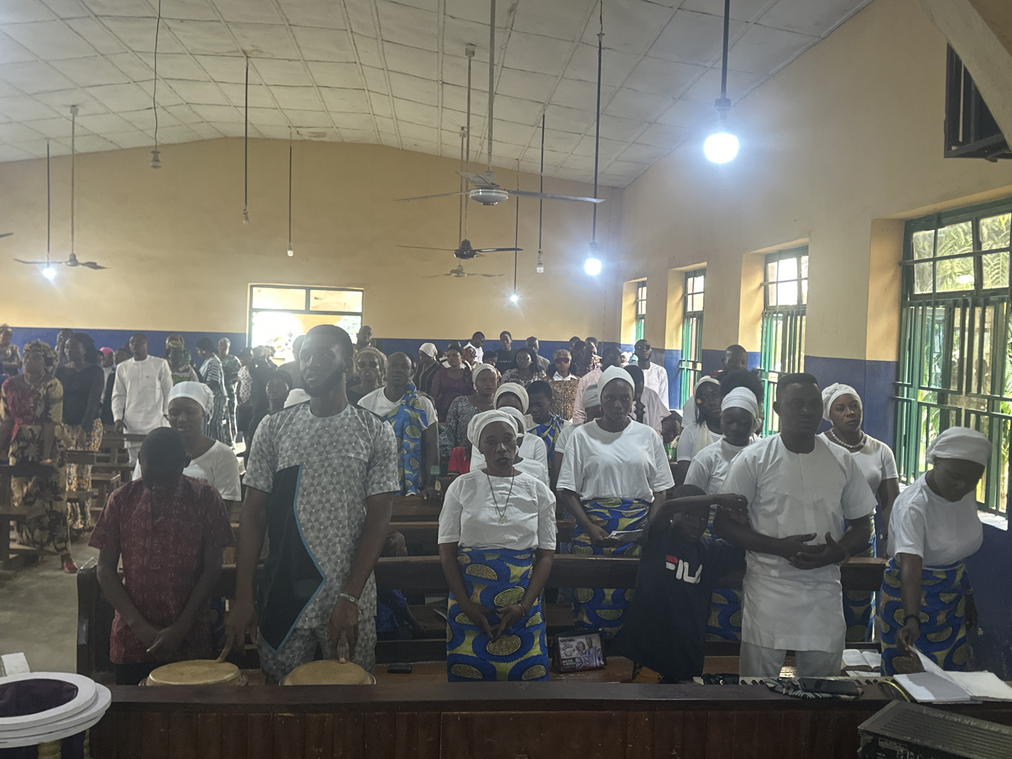 The image depicts a gathering of people, likely a congregation, inside a room with a yellow-painted wall and a white tiled ceiling. Several ceiling fans are visible. The room appears to be a place of worship or a community hall. The people are mostly facing forward, with many wearing white tops and patterned skirts or wrappers. Some individuals are wearing headscarves. In the foreground, there are wooden benches and what appear to be drums. The overall atmosphere seems solemn and participatory. The scene is set in Akwanga, Nigeria.